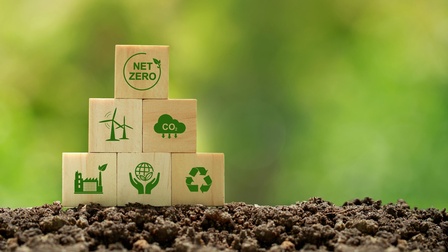 Mehrere Holzwürfel aufeinander gestapelt auf Erde mit grünen Symbolen zum Thema Klimaschutz, oberster Würfel mit Schriftzug Net Zero, Hintergrund verschwommen grün 