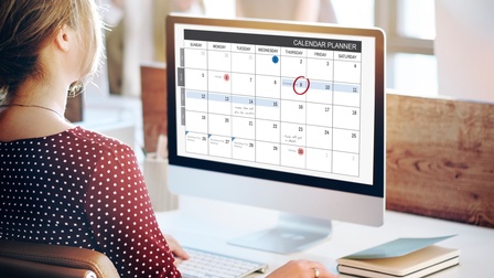 Person in Rückansicht sitzt an Schreibtisch mit Computermonitor, auf dem Kalender zu sehen ist