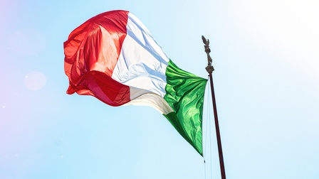 Italienische Flagge, rot-weiß-grün gestreift, an Masten mit Adler aus Metall an der Spitze im Wind wehend vor blauem Himmel