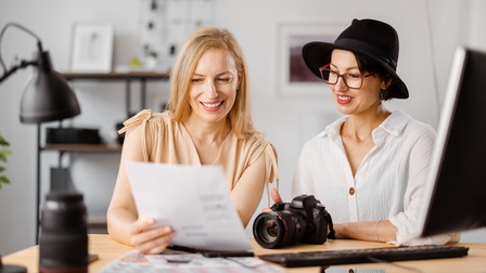 Zwei lächelnde Personen blicken auf Blatt, eine Person trägt Hut und Brille, ringsum Arbeitsmaterialien und Fotoequipment