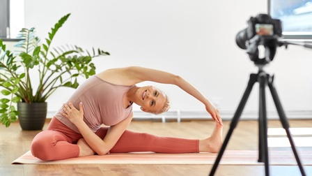 Jugendliche Person in Fitnesskleidung zeichnet Yoga-Übungen mittels einer Kamera auf, die auf einem Stativ steht während sich im Hintergrund ein heller Wohnraum mit einer Grünpflanze befindet