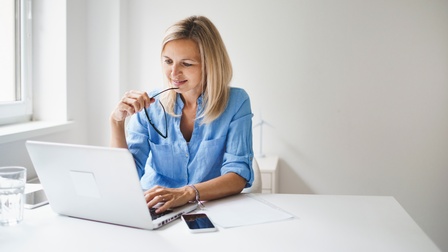 Blonde Person sitzt vor aufgeklapptem Laptop und hält Brillenbügel an die Lippen