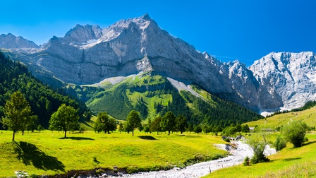 Schroffe Berglandschaft im Sonnenschein unter blauem Himmel umsäumt von bewaldeten Hügeln, im Vordergrund reißender Bach umgeben von sattgrünen Wiesen mit Laubbäumen