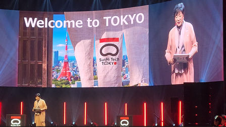 Eine Person steht auf einer Bühne. Hinter der Person ist eine große Leinwand, auf der rechts die Person ist und links daneben Welcome to Tokyo steht