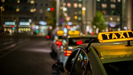 Leuchtendes Zeichen auf Autodach in der Nacht: Gelber länglicher Leuchtkörper mit schwarzen Elementen und Schriftzug Taxi,  im Hintergrund verschwommen weitere parkende Taxis und reflektierende Lichter