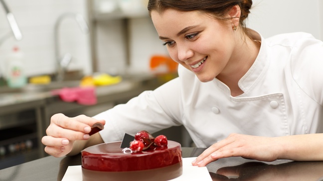 Person mit dunklen geschlossenen Haaren und weißer Arbeitsjacke platziert ein Schokoladeplättchen auf einer roten Torte und blickt freudig darauf, im Hintergrund zeigt sich eine Gastroküche