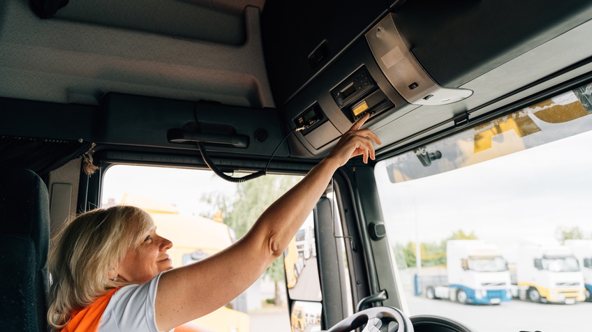 Lächelnde Person in oranger Warnweste sitzt in Fahrerkabine eines Lastkraftwagens und streckt Arm nach oben, um Knopf zu betätigen, im Hintergrund verschwommen weitere parkende Lastkraftwagen