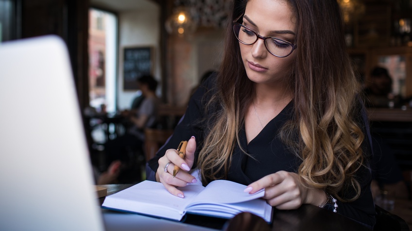Brillen tragende Person mit langen dunklen Haaren blickt in vor ihr liegendes Kalenderbuch und hält Kugelschreiber in einer Hand, im Hintergrund verschwommen Lokal mit weiteren Personen