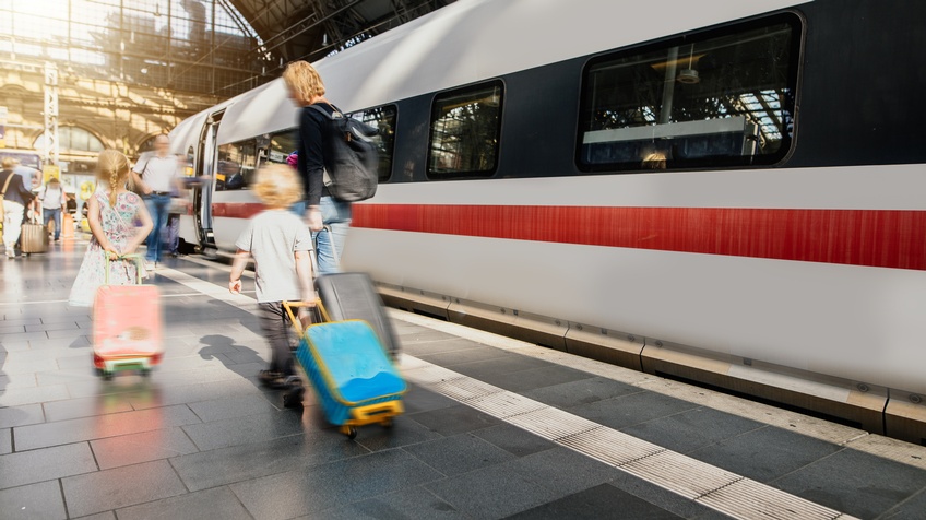Erwachsene Person und zwei Kinder in Bewegungsunschärfe Rollkoffer auf Bahnsteig hinter sich her ziehend, daneben im Ausschnitt weißer Zug mit rotem mittigem Streifen und schwarz eingefassten Fenstern