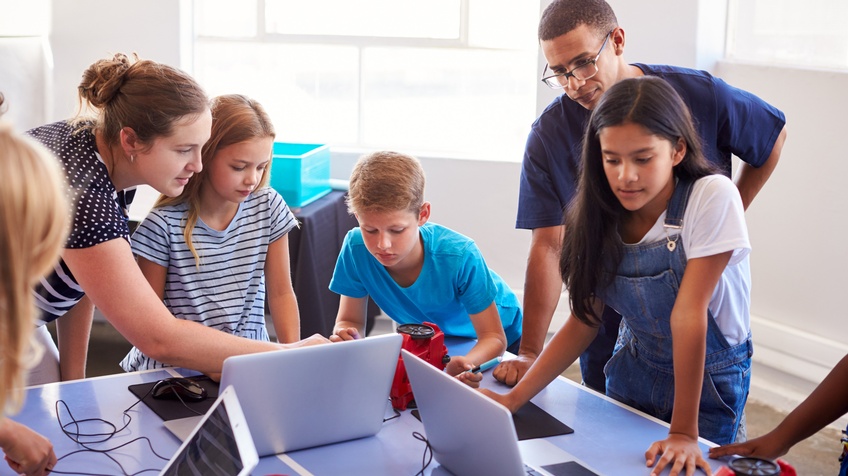 Mehrere Kinder um Tisch versammelt, auf dem Laptops stehen, zwei Erwachsene lehnen über Tisch