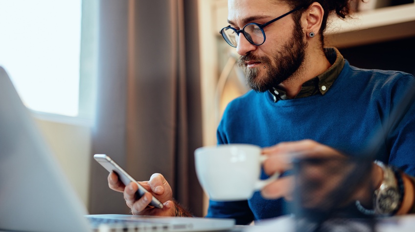 Bärtige Person mit Brille hält in einer Hand Tasse, in anderer Hand Smartphone, daneben aufgeklapptes Notebook