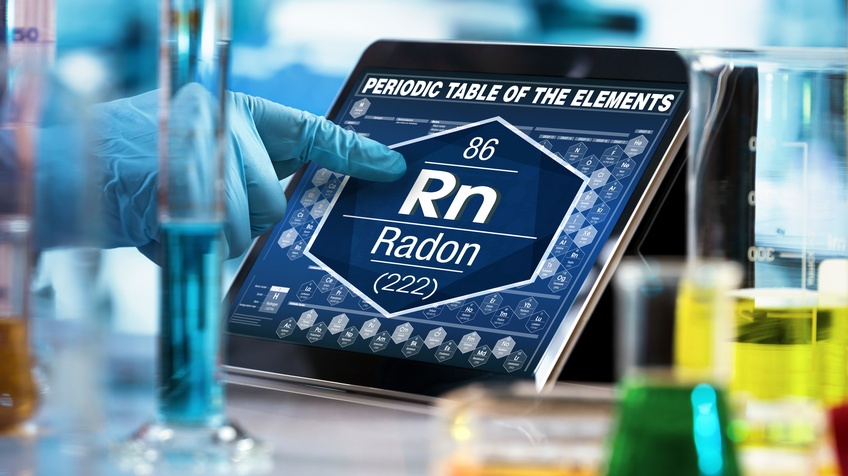 Fokus auf ein Tablet auf dessen Bildschirm Radon aus dem periodischen System aufscheint, eine Hand in Gummihandschuh deutet darauf, ringsum Phiolen mit verschiedenfarbigen Flüssigkeiten.