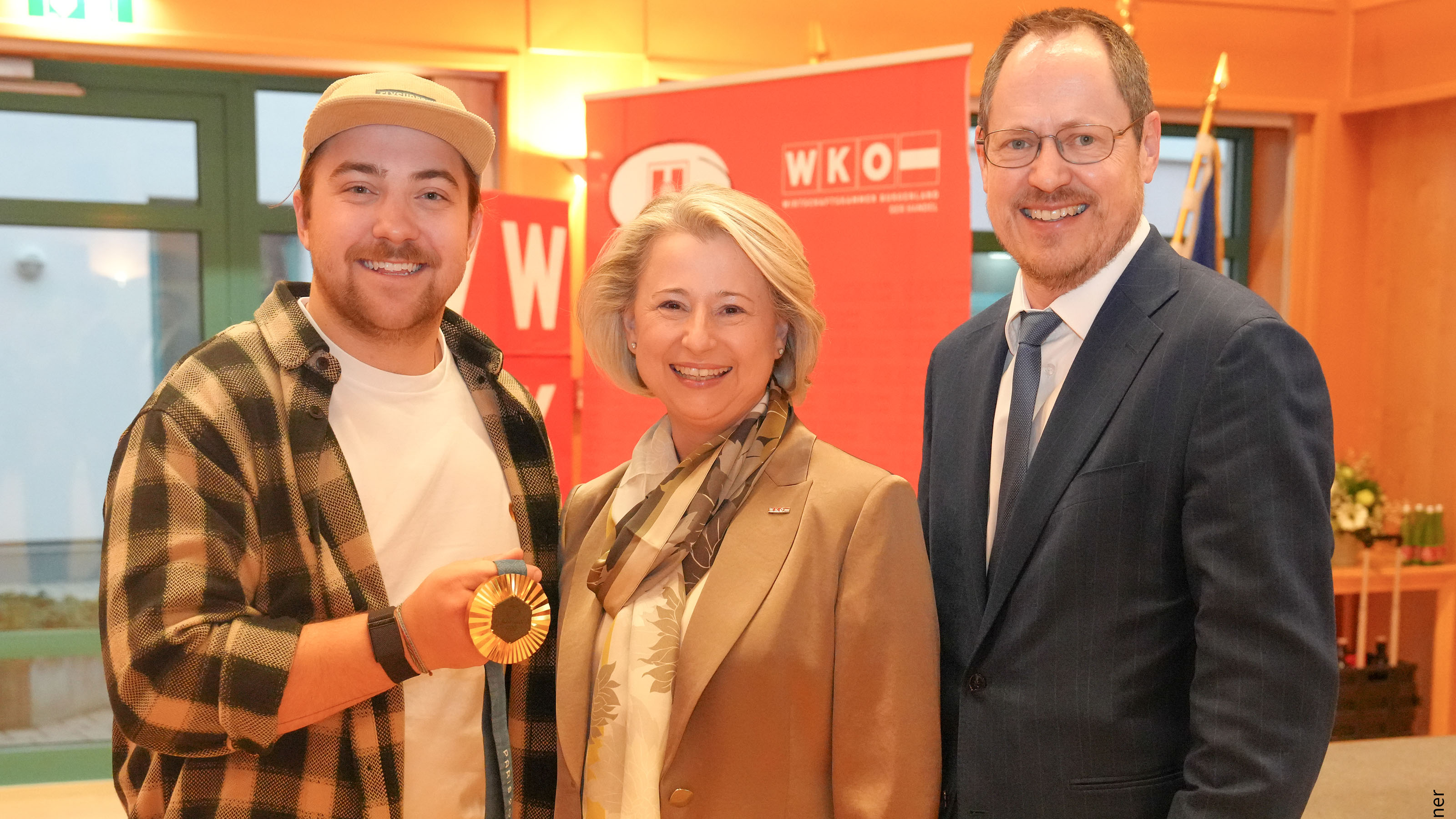 Drei Personen - der Mann ganz links hält eine Gold-Medaille in die Kamera