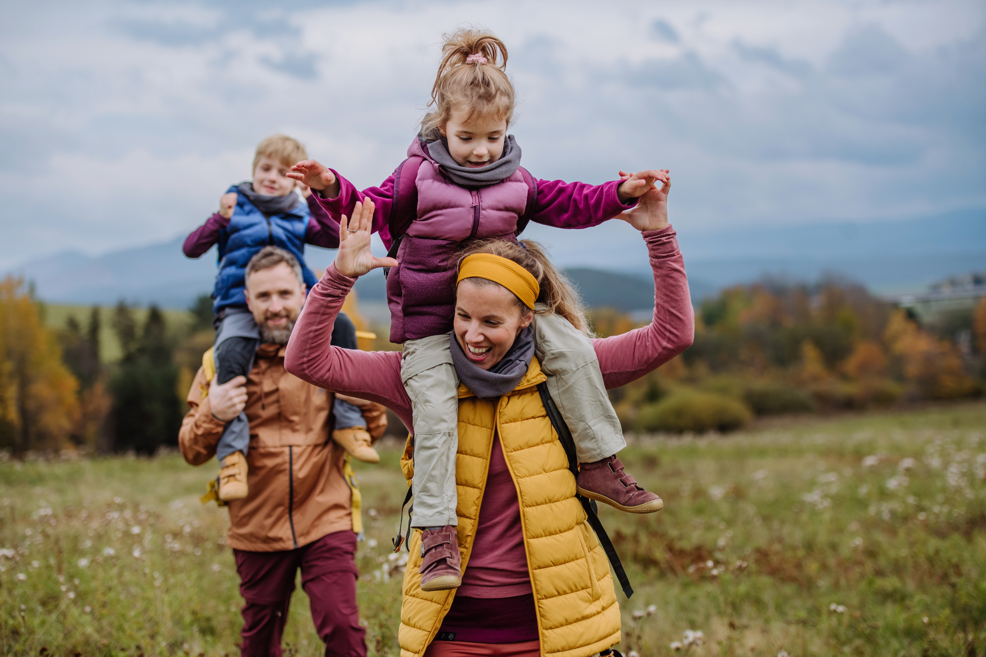 Zwei Erwachsene und zwei Kinder in Wanderkleidung gehen über eine Wiese. Je eine erwachsene Person trägt ein Kind auf ihren Schultern