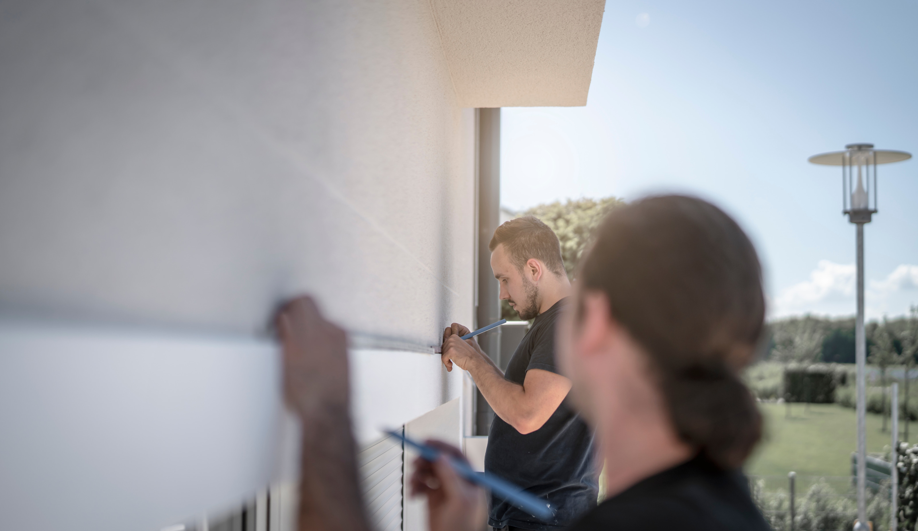 Person im Fokus, die an Hausfassade Paneel für Sonnenschutz anzeichnet, im Vordergrund in Rückenansicht verschwommen weitere Person, im Hintergrund verschwommen Garten und Außenleuchte