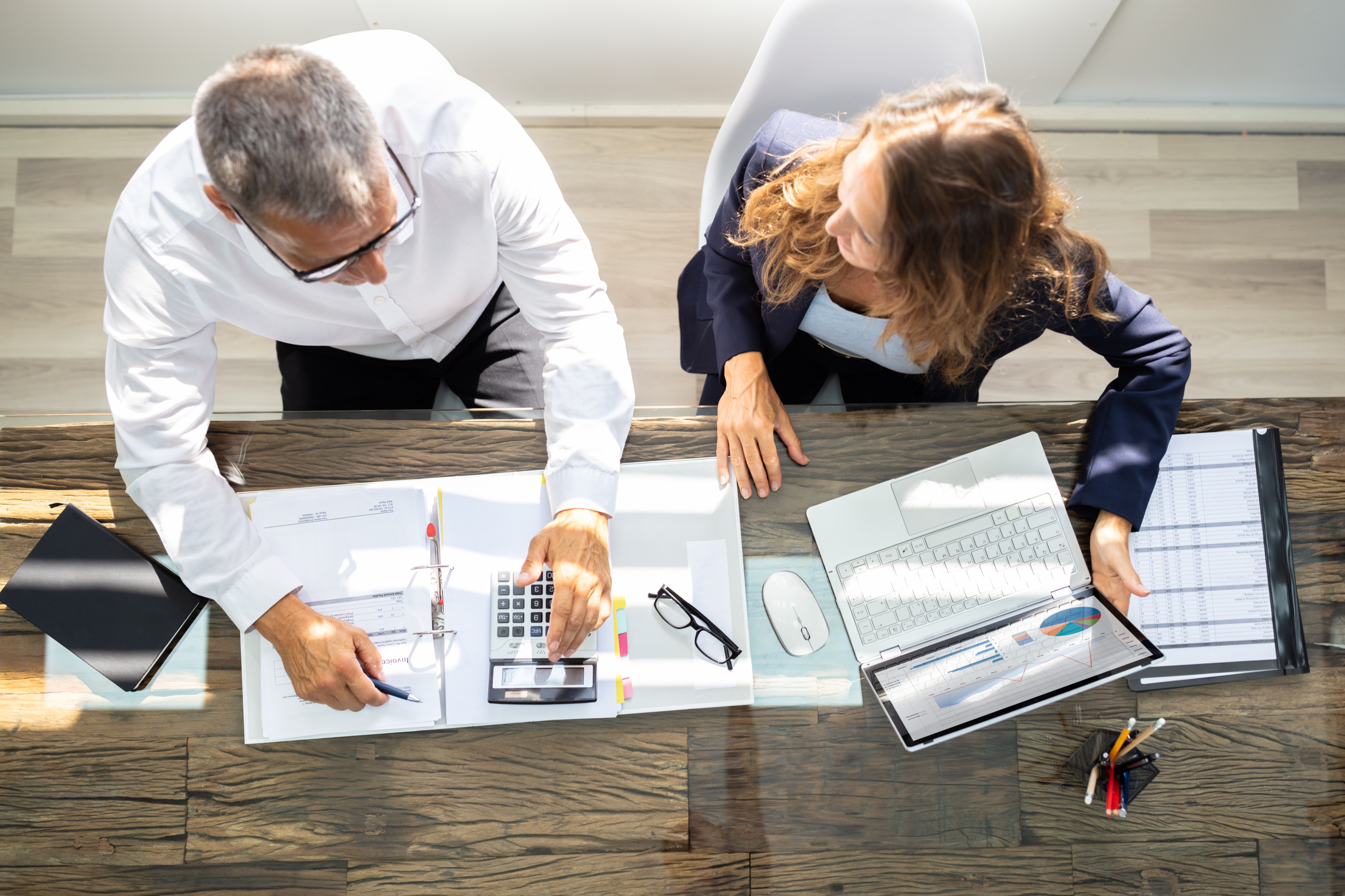 Zwei Personen in Businesskleidung halten ein Meeting an einem Schreibtisch mit einem Laptop und weiteren Arbeitsunterlagen wie Mappen, Taschenrechner, Listen, Notizbücher, Brillen und Stiften, Topview