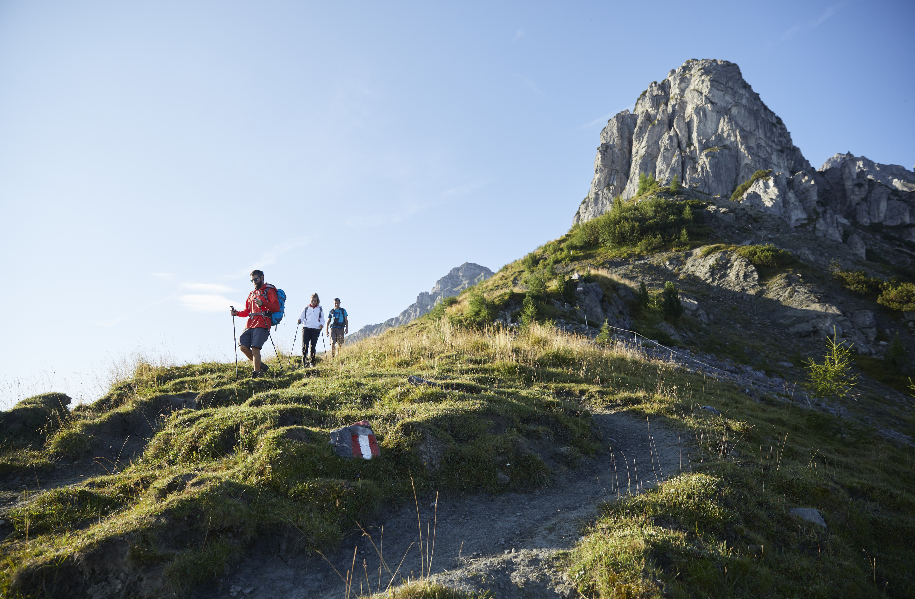 Drei Personen in Wanderkleidung, zwei davon mit Wanderstöcken ausgerüstet, marschieren Berg herunter