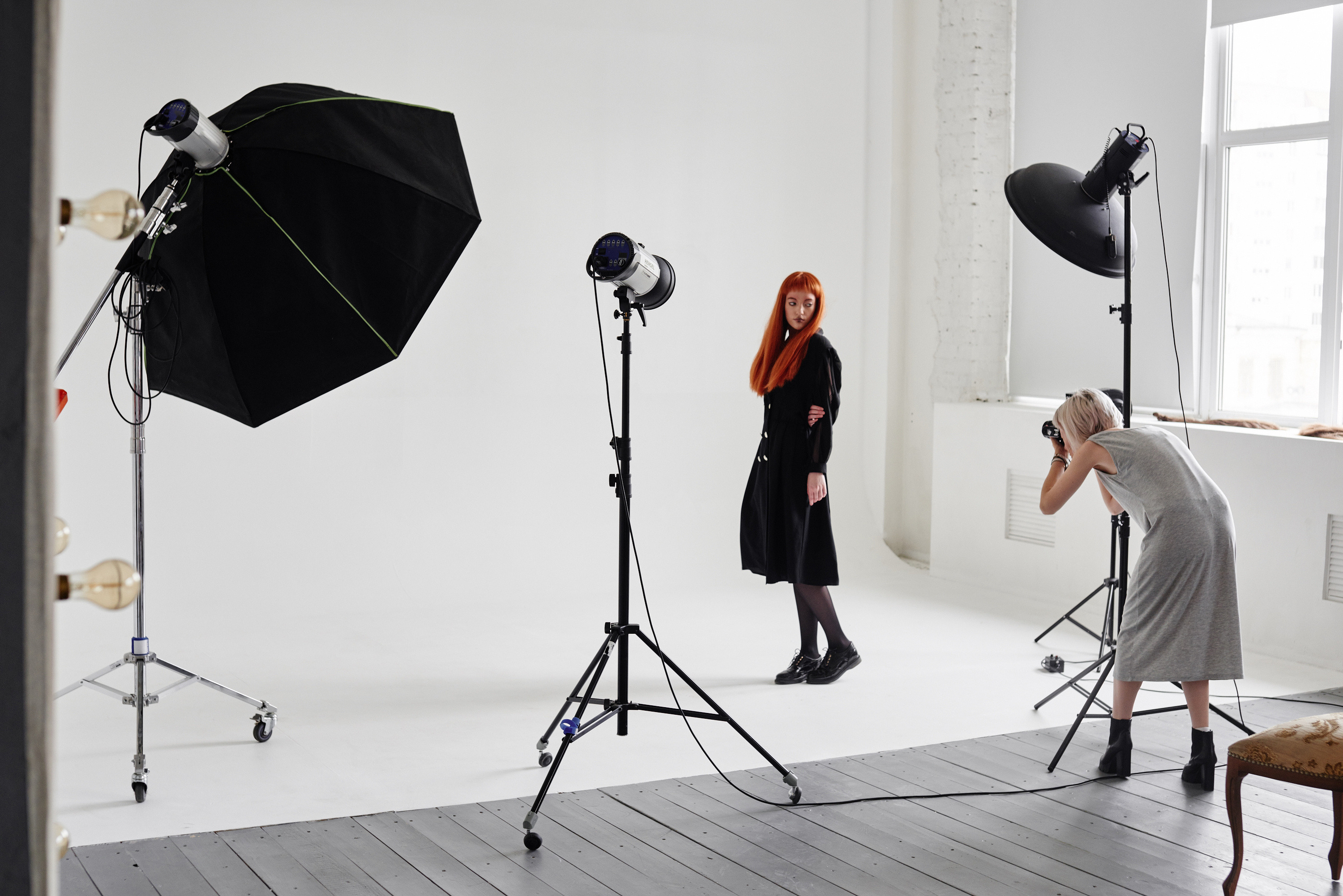 Person in dunklem Kleid und mit langen roten Haaren posiert vor weißem Hintergrund für andere Person, die Fotos mit Kamera macht, ringsum Studioblitze