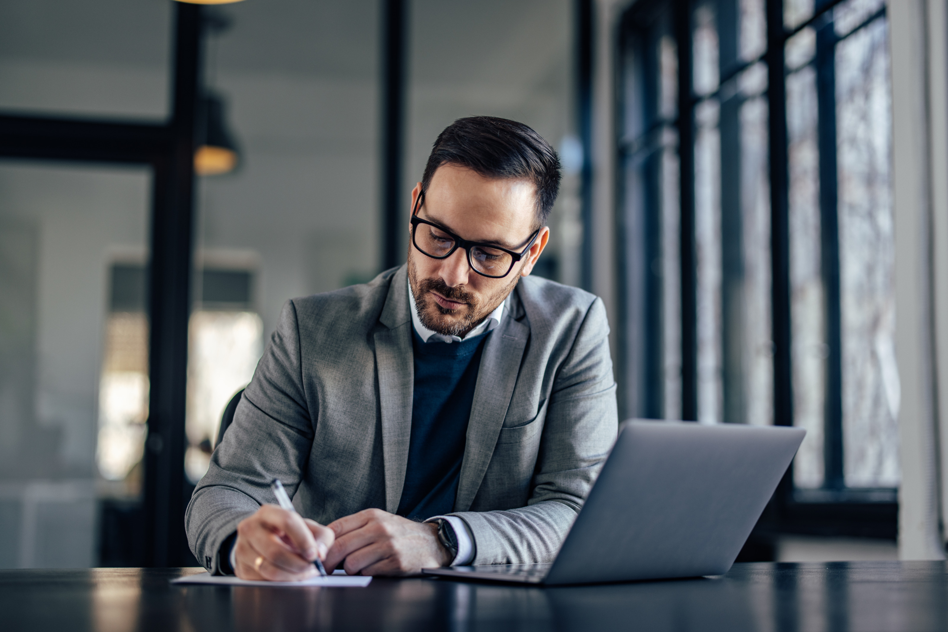 Person mit kurzen Haaren, Brille und Bart trägt Businesskleidung und arbeitet bei einem Schreibtisch mit einem Laptop in einem Büro