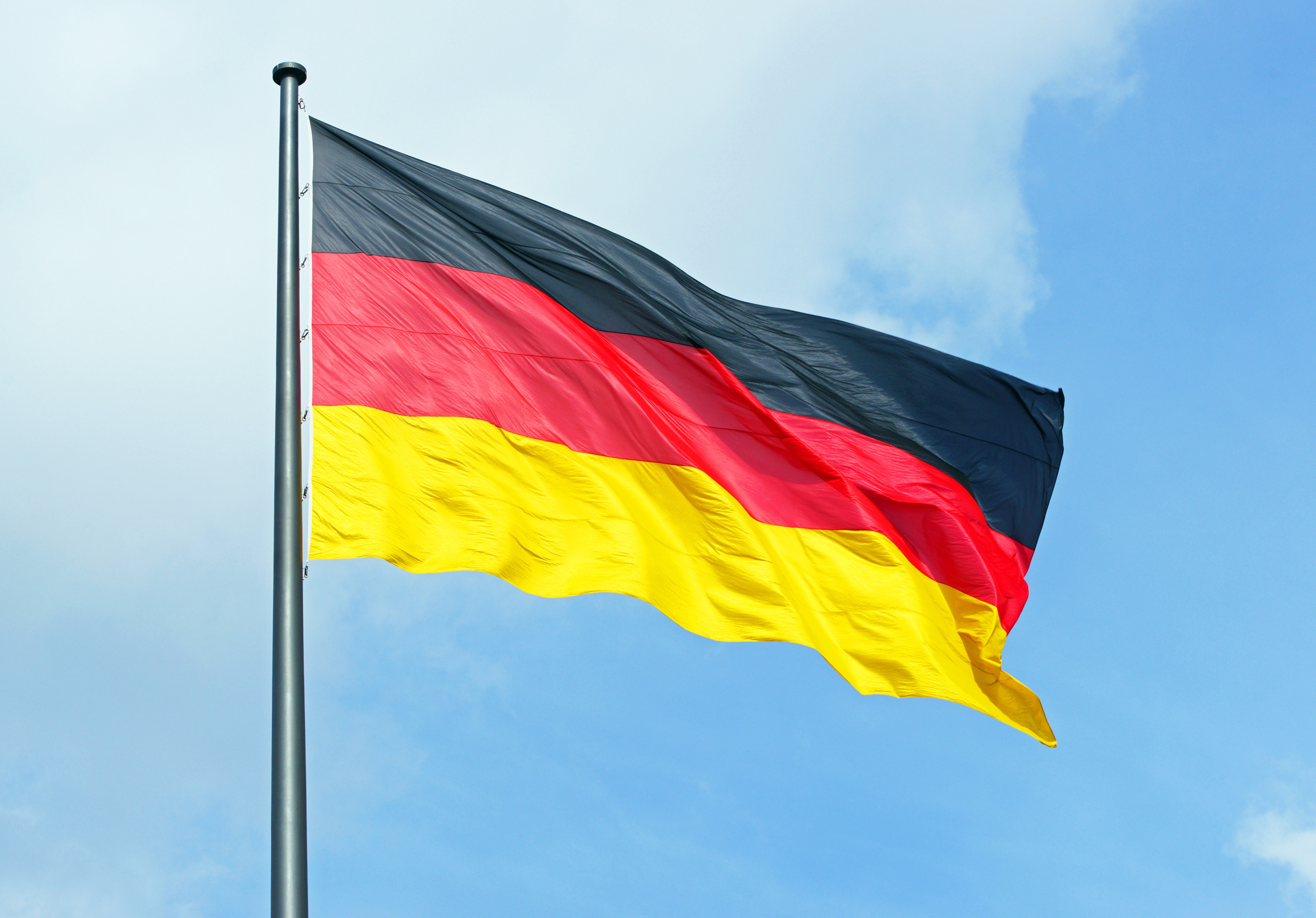 An einer Metallstange ist die Flagge Deutschlands montiert. Die Flagge weht im Wind. Dahinter ist der blaue Himmel mit einigen weißen Wolken