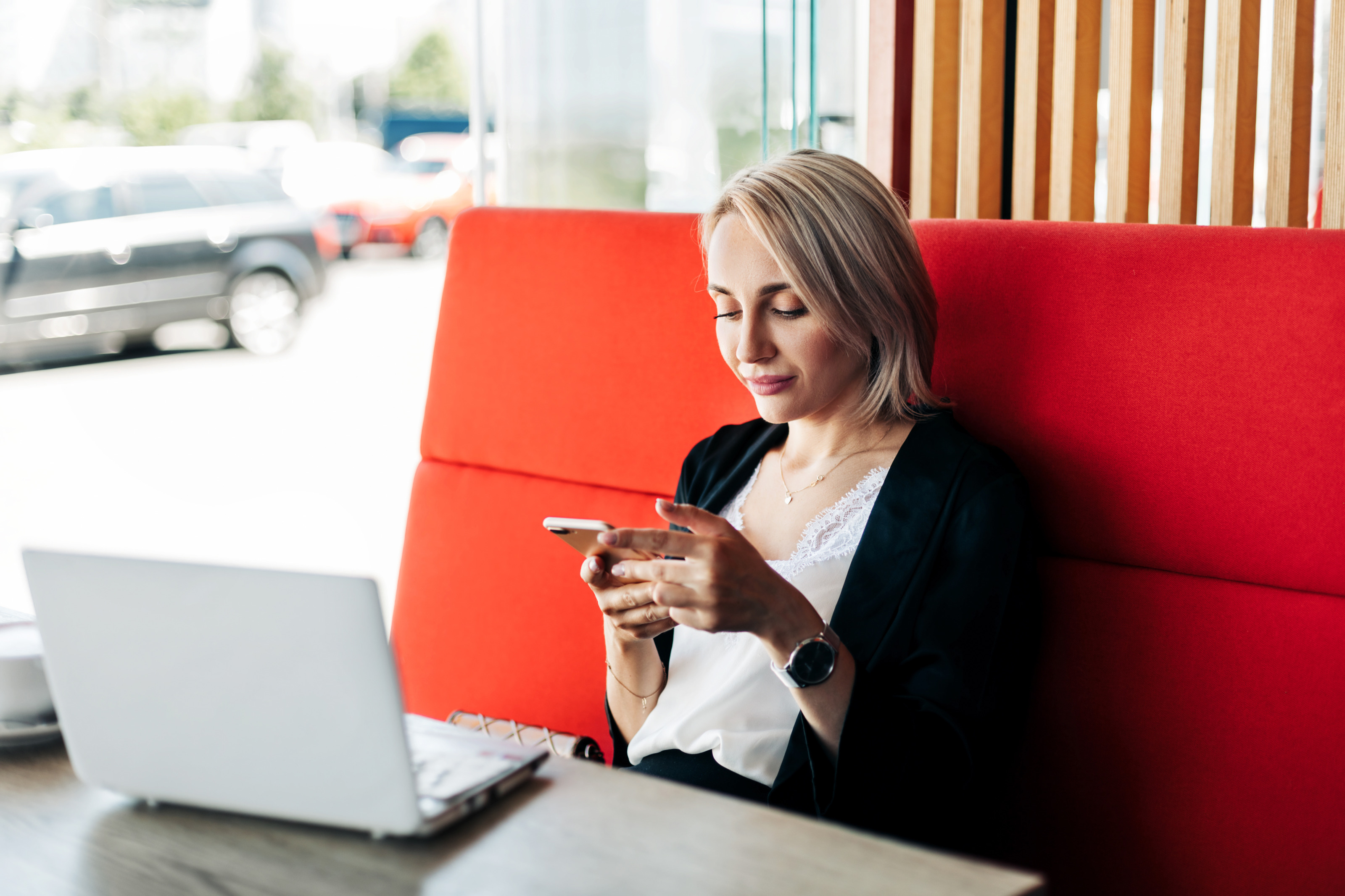 Person mit blonden Haaren sitzt  bei einem Tisch in einem Restaurant und blickt auf ein Smartphone während ein Laptop und eine Tasse auf dem Tisch stehen