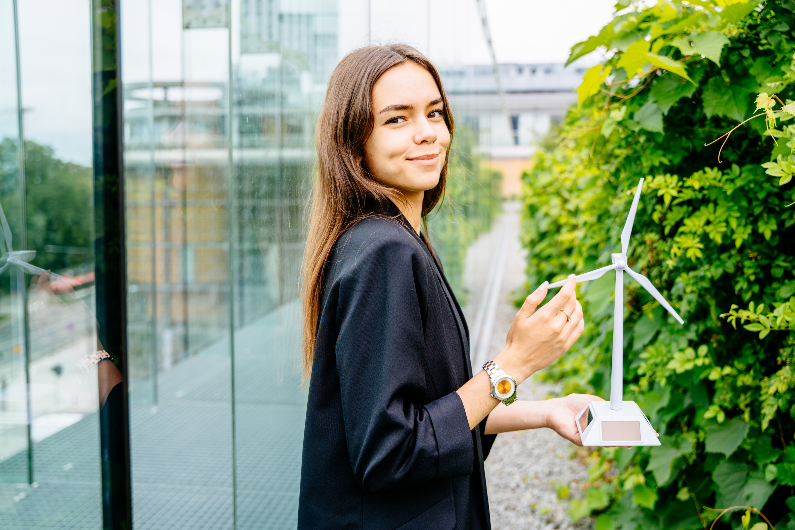 Sanft lächelnde Person hält Modell eines Windrades in Händen, zwischen Glasfassade eines Gebäudes und grüner Hecke stehend