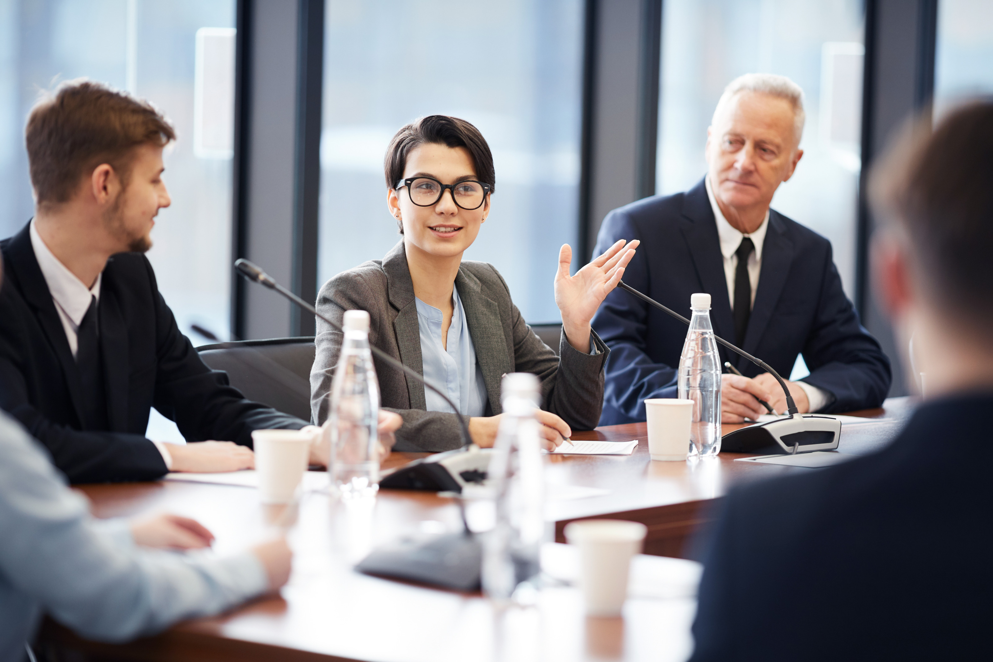 Person mit kurzen dunklen Haaren, Brille und Businesskleidung sitzt während einer Konferenz am Tisch mit mehreren anderen Personen, die aufmerksam auf die sprechende Person blicken