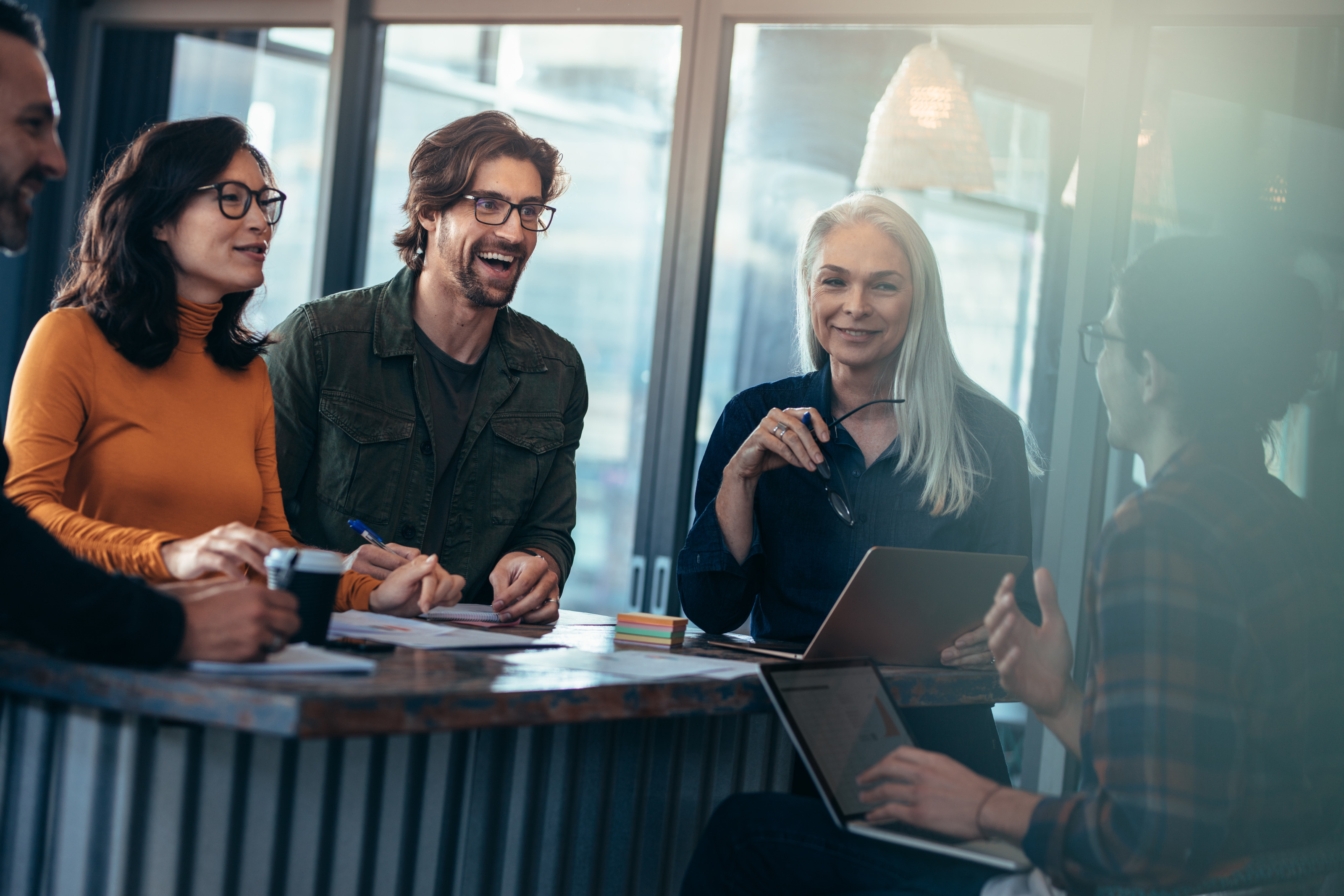 Personen stehen und sitzen freudig rund um einen Tisch bei einem Meeting mit Unterlagen und Laptops