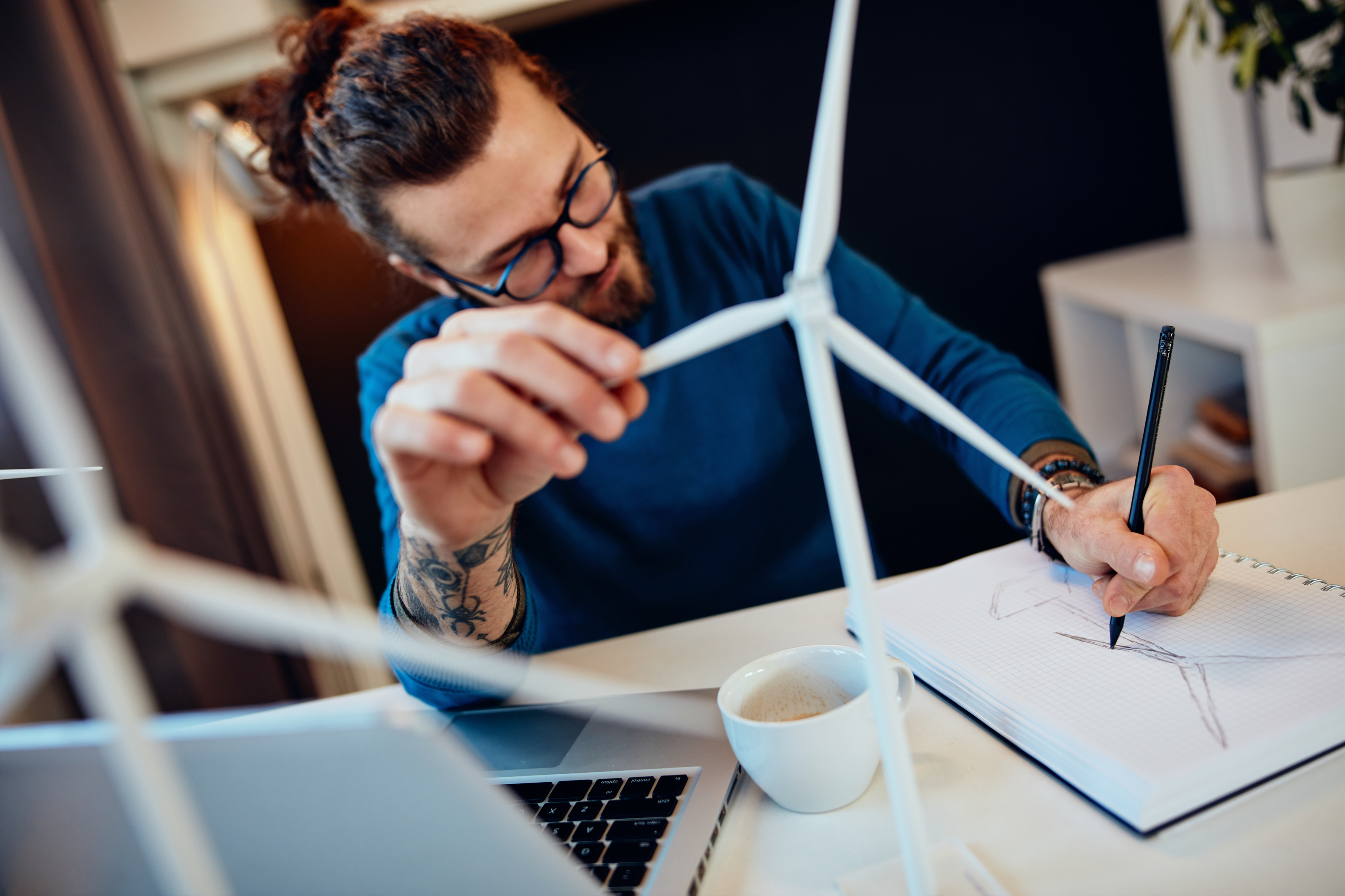 Person mit Brillen sitzt an Schreibtisch, skizziert Windrad auf Block und hält in einer Hand Modell eines Windrades, am Tisch aufgeklappter Laptop und Kaffeetasse