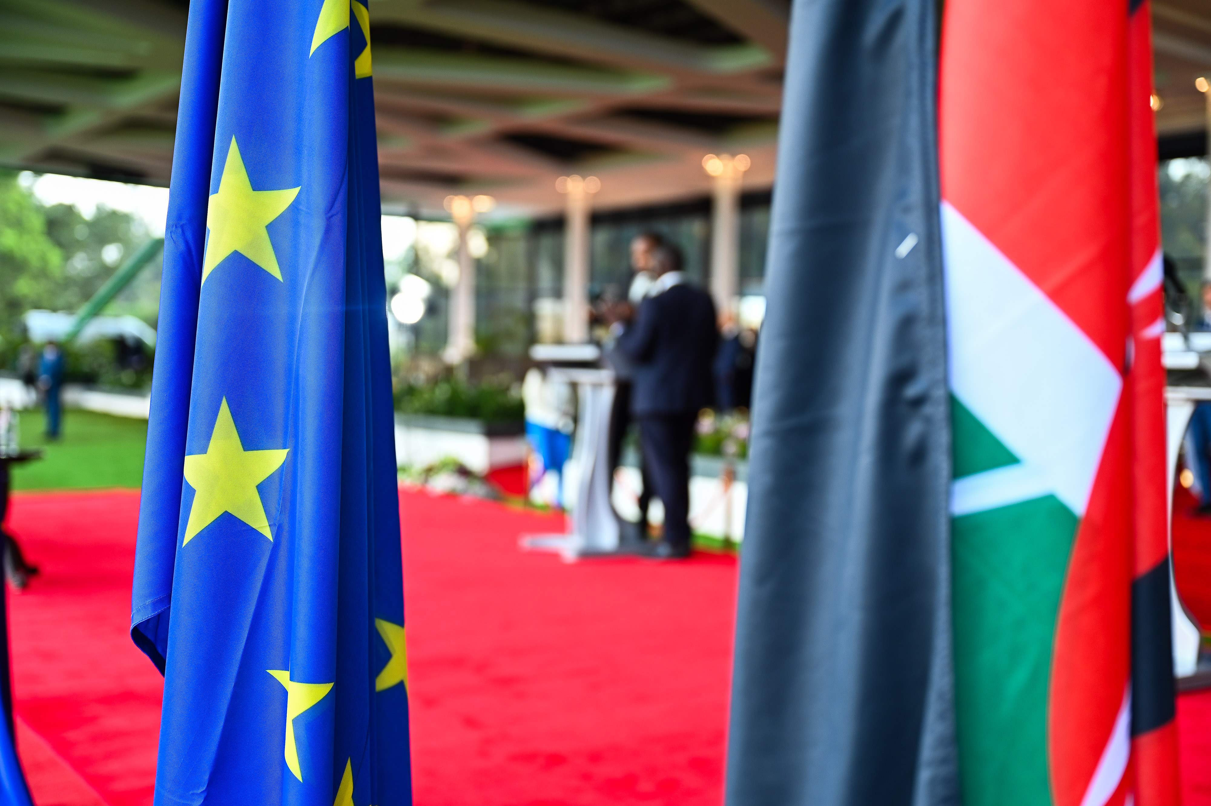 Im Vordergrund ist links die Flagge der Europäischen Union. Rechts ist die Flagge des Landes Kenia. Im Hintergrund stehen zwei Personen auf einem roten Teppich