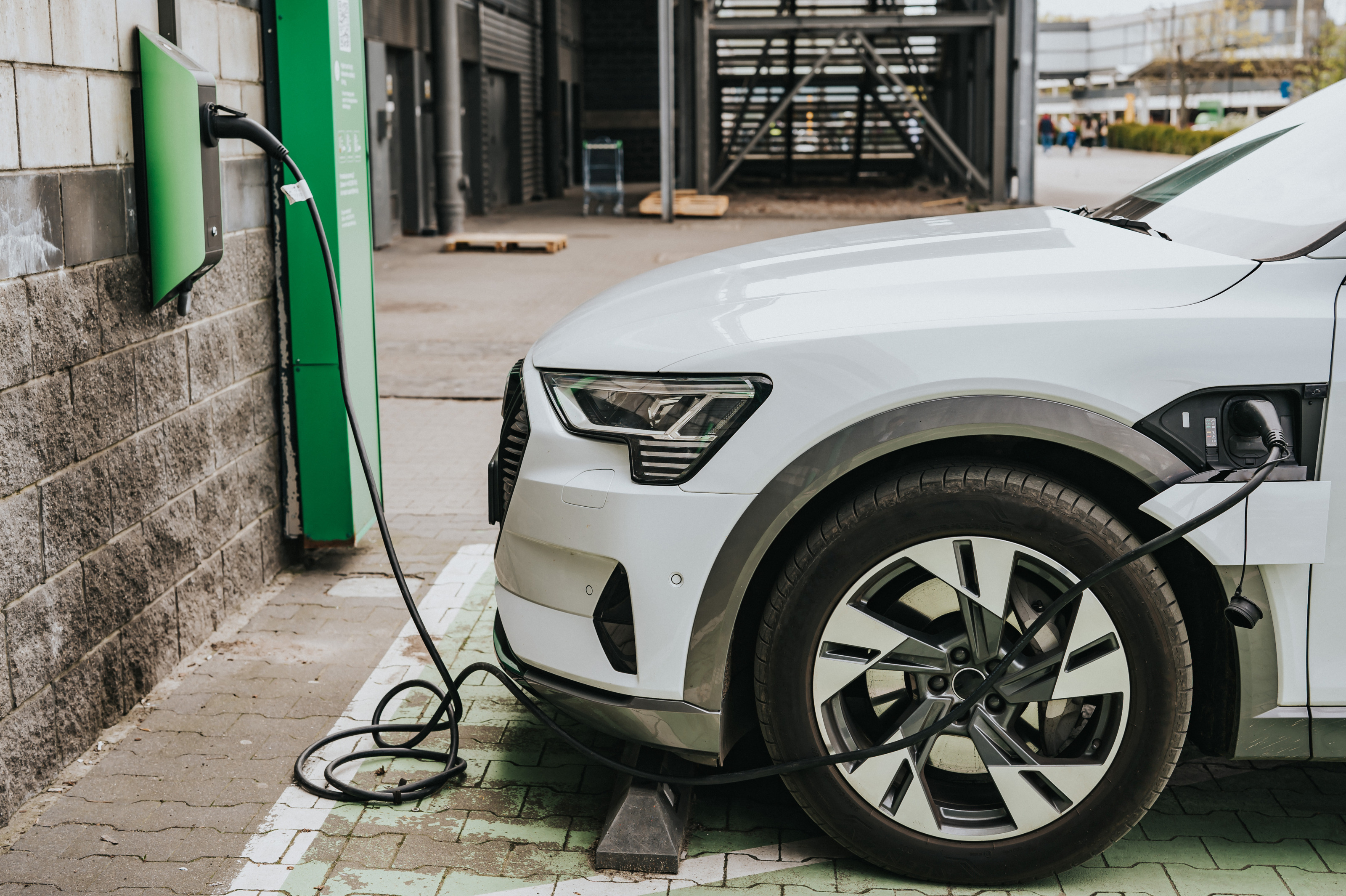 Ein weißes Elektroauto ist zur Hälfte zu sehen. Es wird an einer E-Tankstelle aufgeladen, die durch ihre grüne Farbe das Bild dominiert. Die Ladestation befindet sich an einer Steinmauer.  Im Hintergrund sieht man verschwommen eine  Garage, 