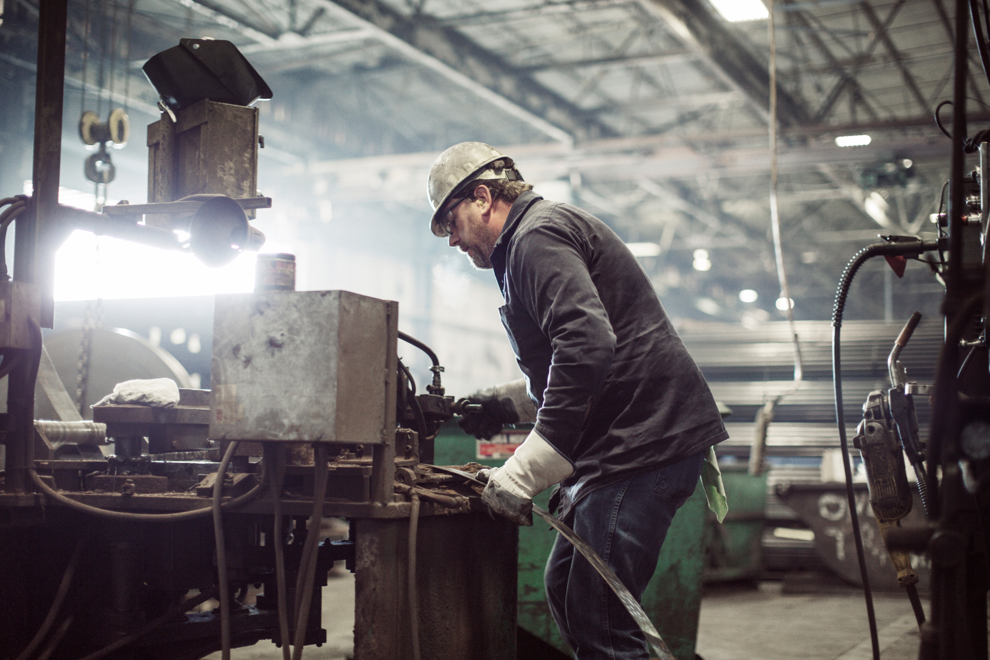 Person mit Schutzhelm und Schutzhandschuhen steht in großer Werkhalle an  Maschine und hält Metallblatt unter Gerät