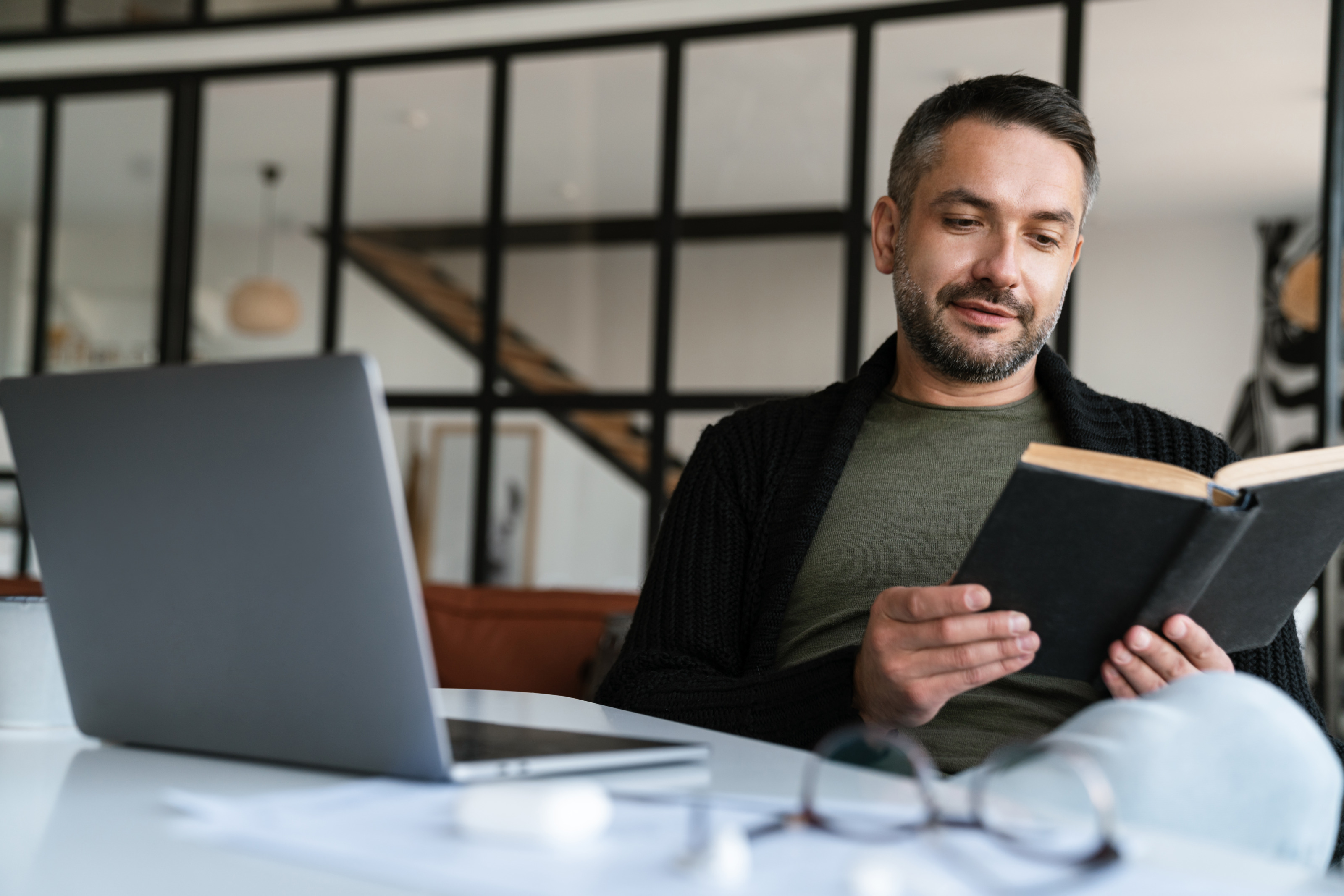 Ein Mann liest ein Buch mit schwarzem Einband. Er sitzt in seiner Wohnung, die wie ein Loft aussieht und im Hintergrund zu sehen ist. Der Laptop ist aufgeklappt, seine Brille ist abgelegt und liegt am Tisch, sie ist verschwommen zu sehen. 