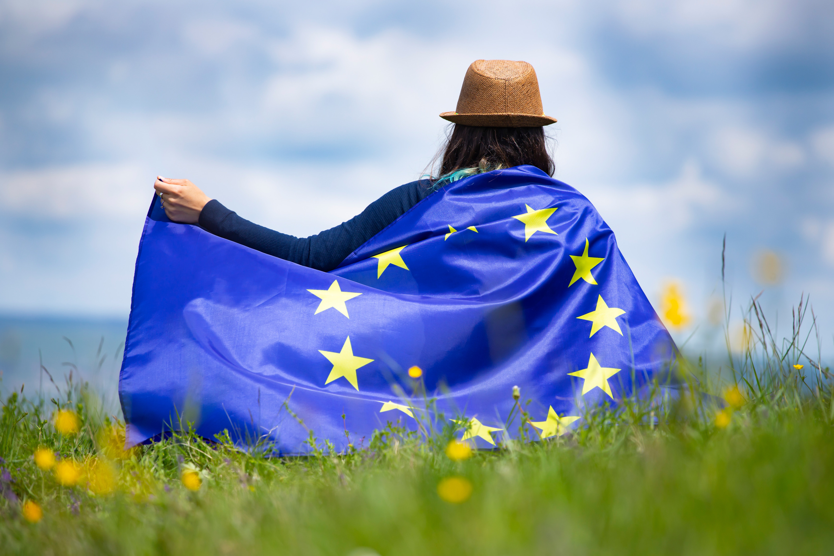 Person in Rückenansicht mit Hut und Europaflagge um die Schultern: Blauer Stoff mit im Kreis verlaufenden gelben Sternen. Die Person wandert über sommerliche Wiese und streckt einen Arm vom Körper, in einer Hand die Ecke der Flagge haltend.