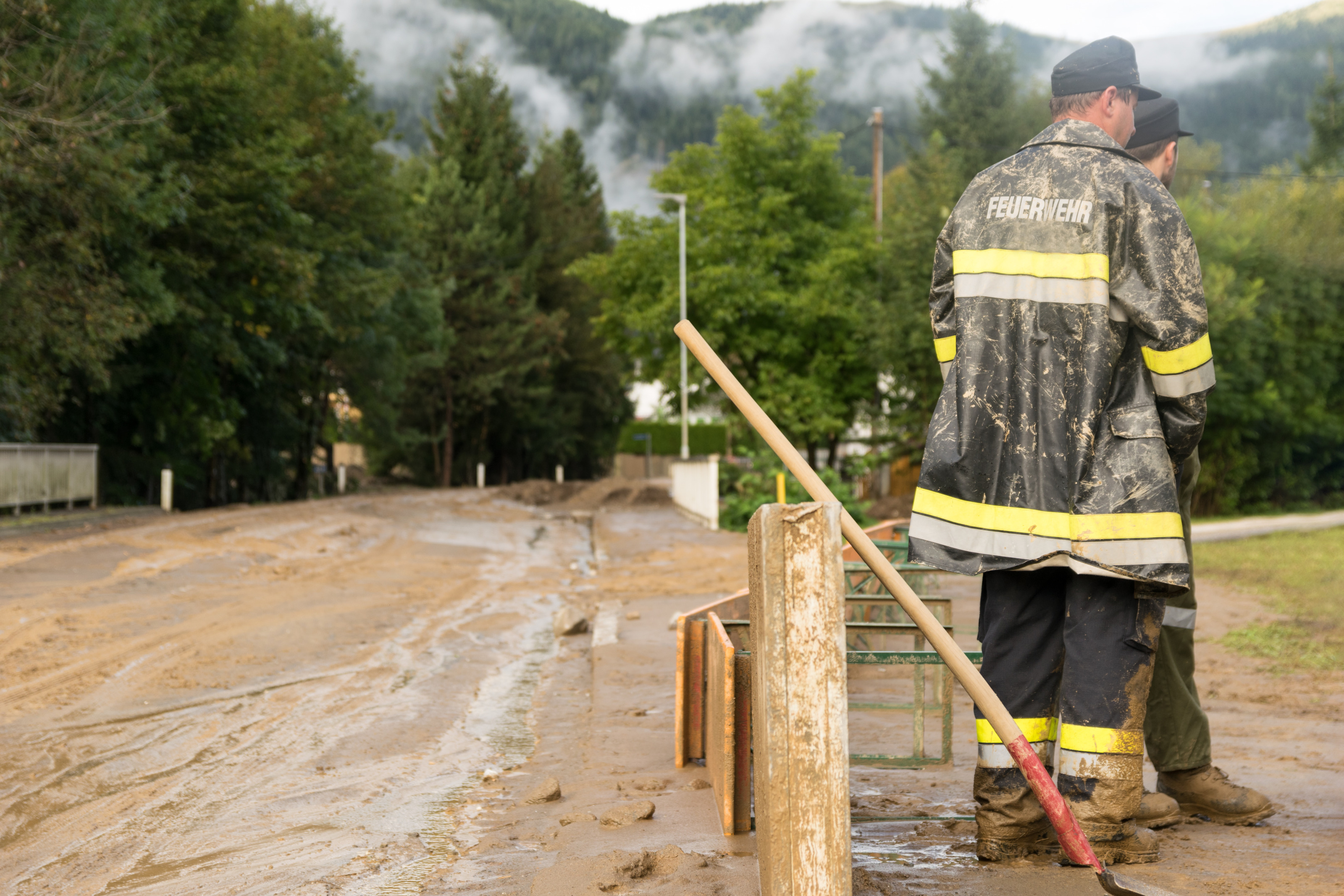 Zwei Personen in Rückenansicht in Schutzkleidung mit der Aufschrift Feuerwehr stehen neben wasserverschlammter Straße, die von Bäumen umsäumt ist, neben den Personen lehnt eine Schaufel an Holzbalken.