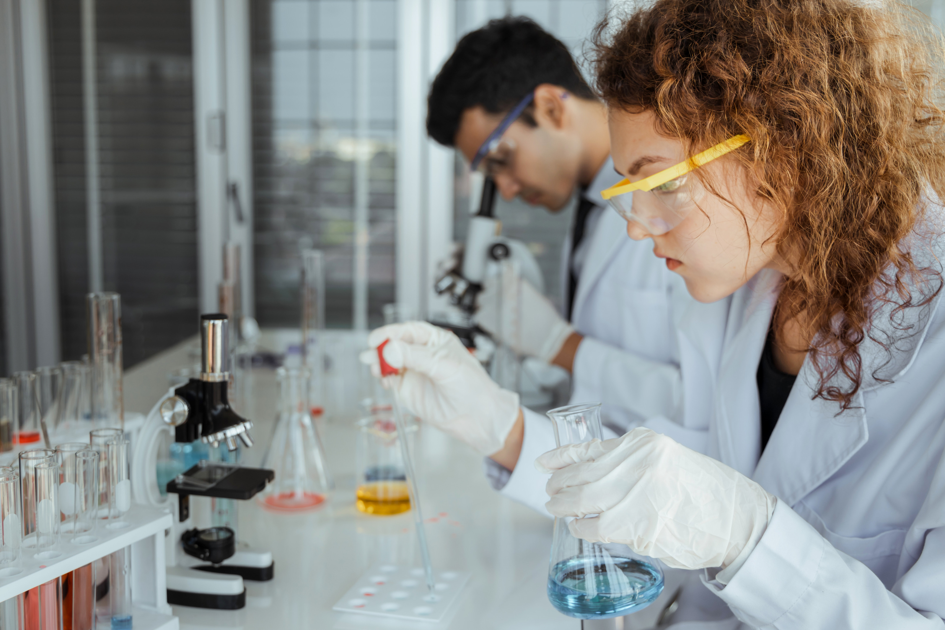 Person mit Schutzbrillen in weißem Kittel an Tisch sitzend, mit Pipette Flüssigkeit in Schälchen tropfend hält in Hand Glas mit Flüssigkeit, am Tisch Mikroskop und Röhrchen mit Flüssigkeiten, im Hintergrund weitere Person durch Mikroskop äugend