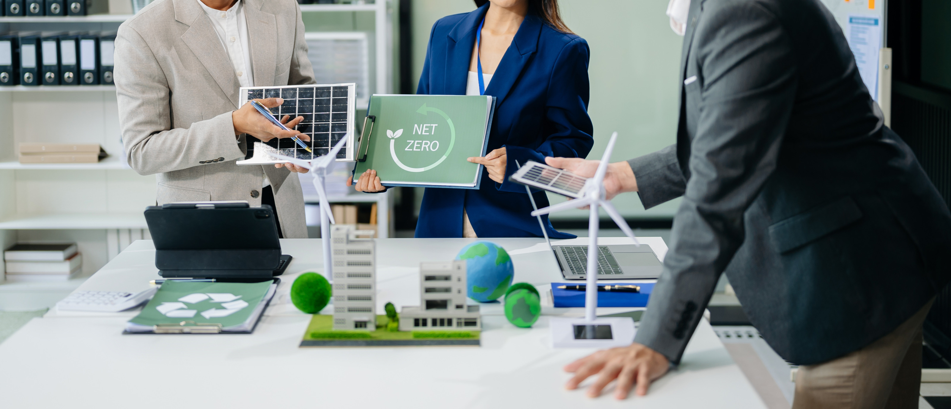 Mehrere Personen im Ausschnitt, nur Oberkörper und Hände ersichtlich, an Tisch stehend mit Modellen wie das eines Windrades, eine Person hält Schild mit Aufschrift Net Zero in Händen