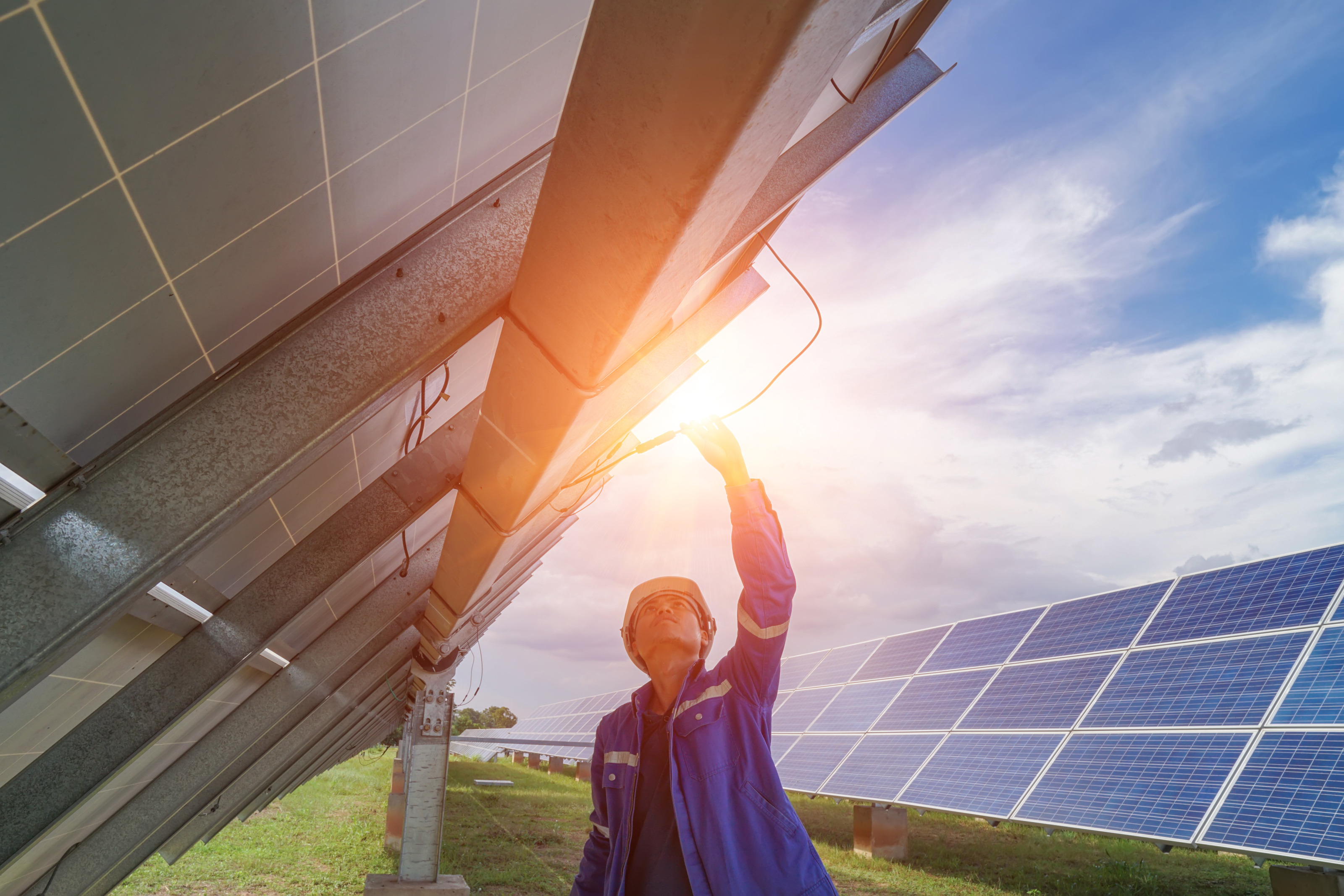 Person mit Helm streckt sich nach Oben und beäugt Kabel einer Solaranlage auf Metallgerüst, im Hintergrund weitere Solarpanele auf grüner Wiese unter wolkenverhangenem Himmel im Sonnenuntergang