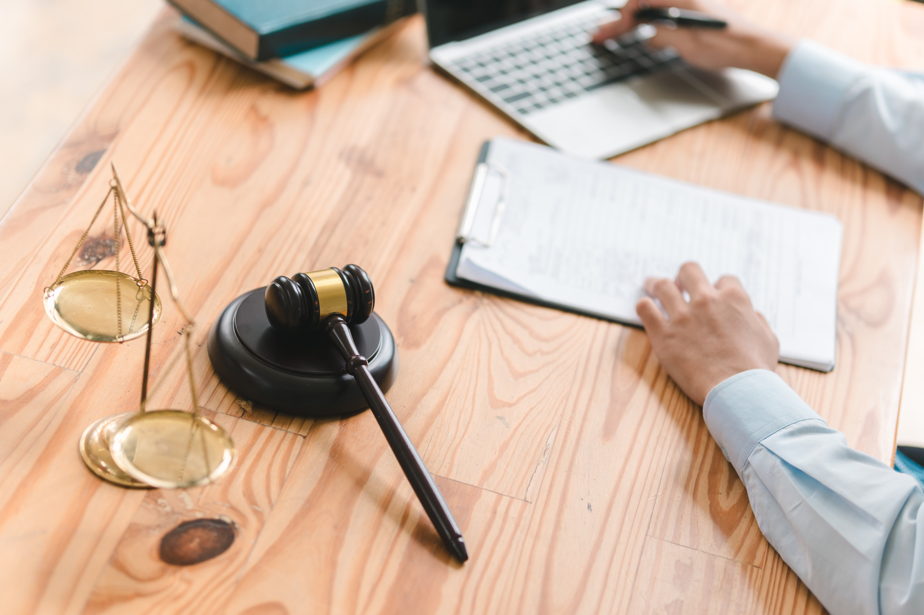 Detailansicht eines Schreibtischs mit Waage und Richterhammer, im Hintergrund verschwommen Hände einer Person, die auf Clipboard und Laptop liegen