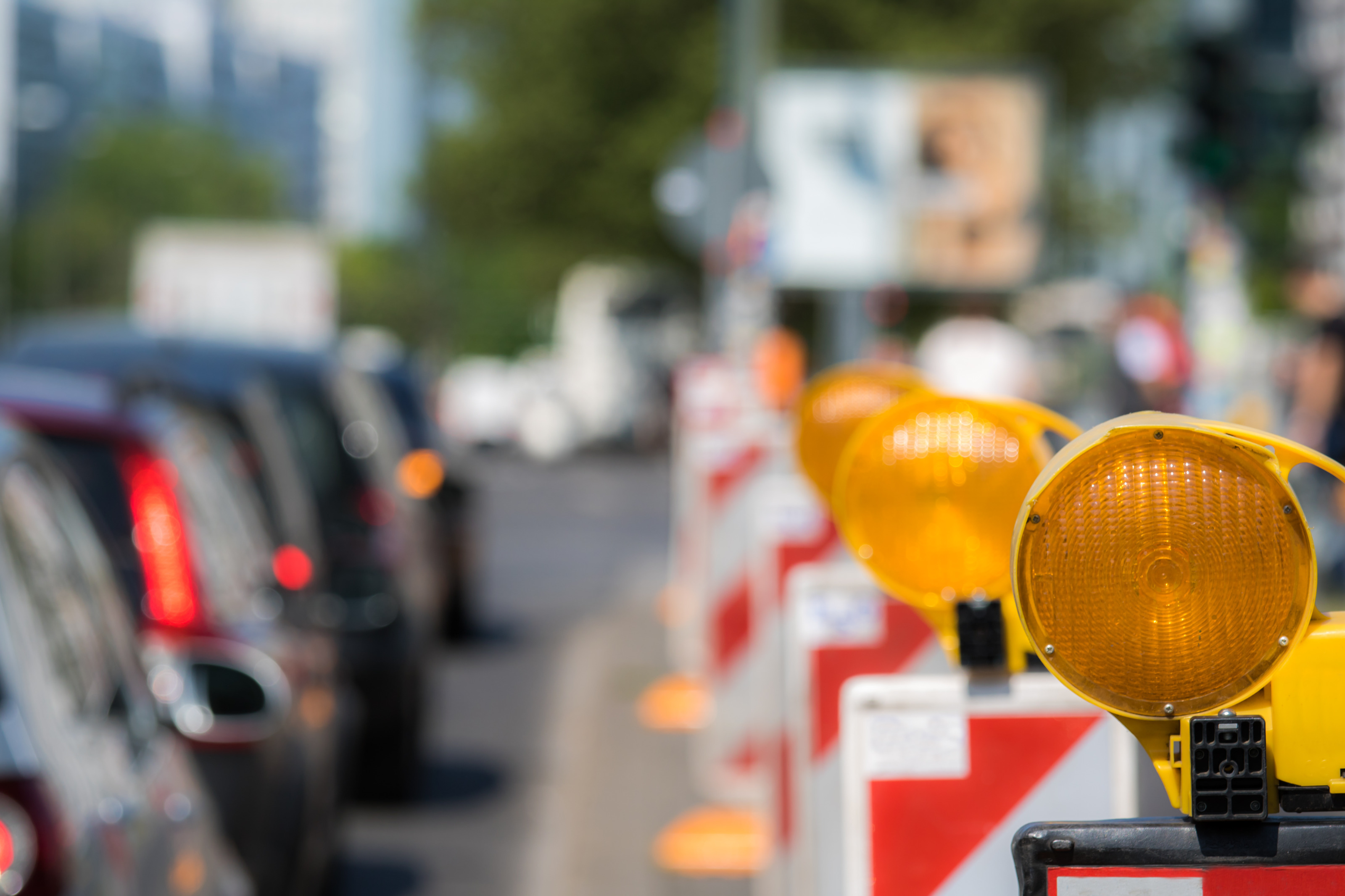 In der linken Bildhälfte sind mehrere hintereinander stehende Autos. In der rechten Bildhälfte sind viele hintereinander stehende rot-weiße Absperrungen. Auf jeder Absperrung ist oben ein oranges Licht montiert.