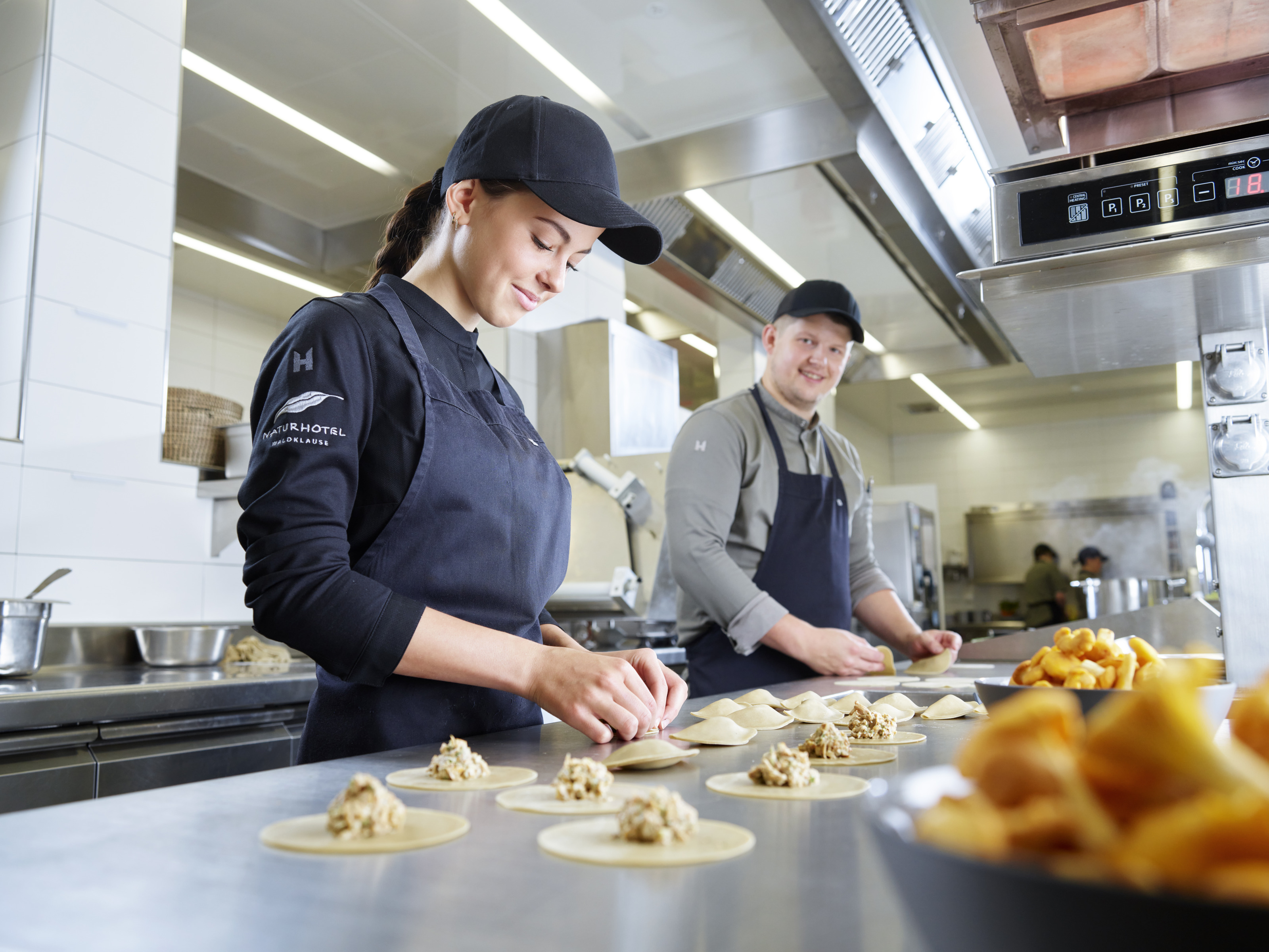 Zwei sanft lächelnde Personen mit Caps und in Schürzen stehen in großer Edelstahlküche und befüllen Teigtaschen, ringsum Küchenutensilien und Schüsseln mit Eierschwammerl