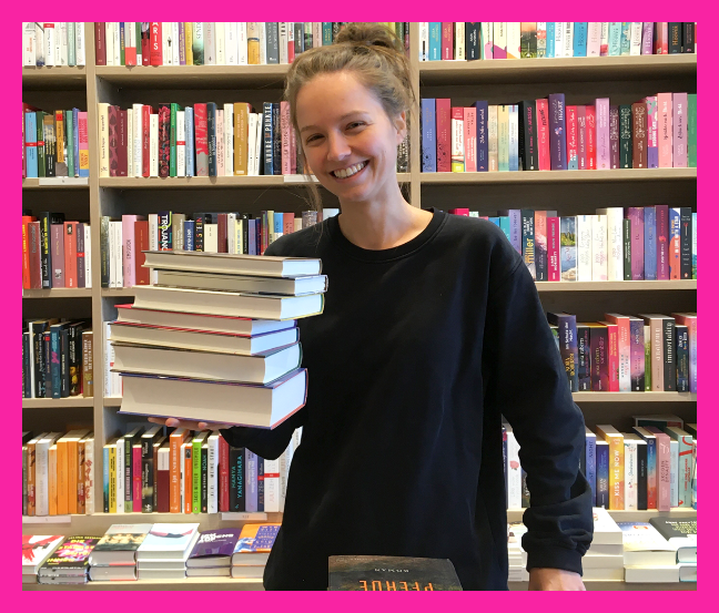 Eine junge Frau lächelt ins Bild und hält einen Bücherstapel in der Hand, im Hintergrund sind Bücherregale zu sehen