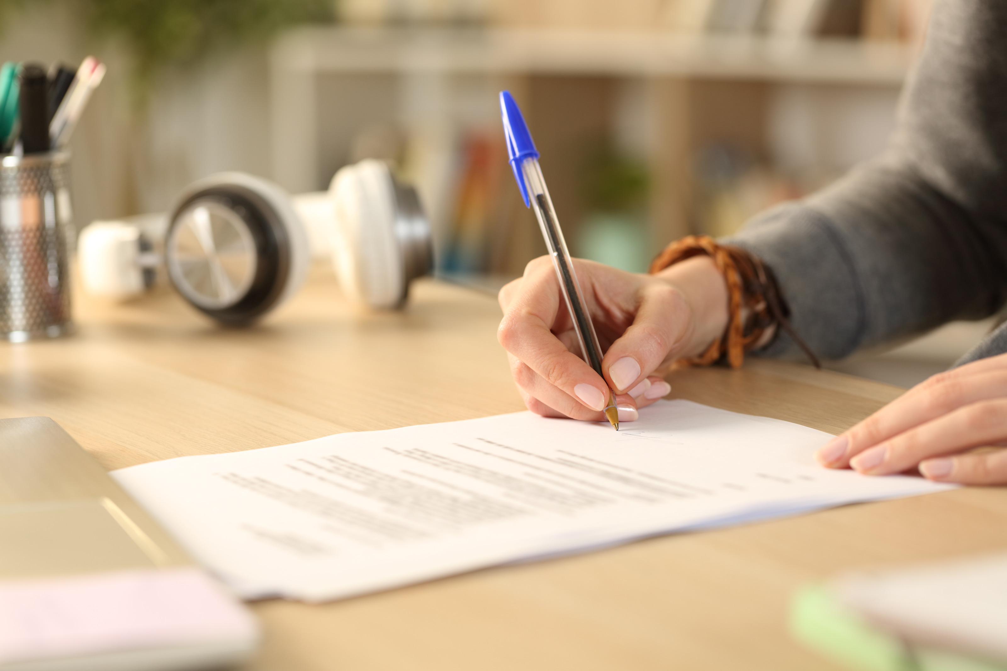 Auf einem Tisch liegen Papiere. Das oberste Blatt ist mit Text beschriftet. Die Hände einer Person liegen auf dem Blatt Papier. Die rechte Hand der Person hält einen Stift und schreibt damit etwas auf die untere Hälfte des Blattes