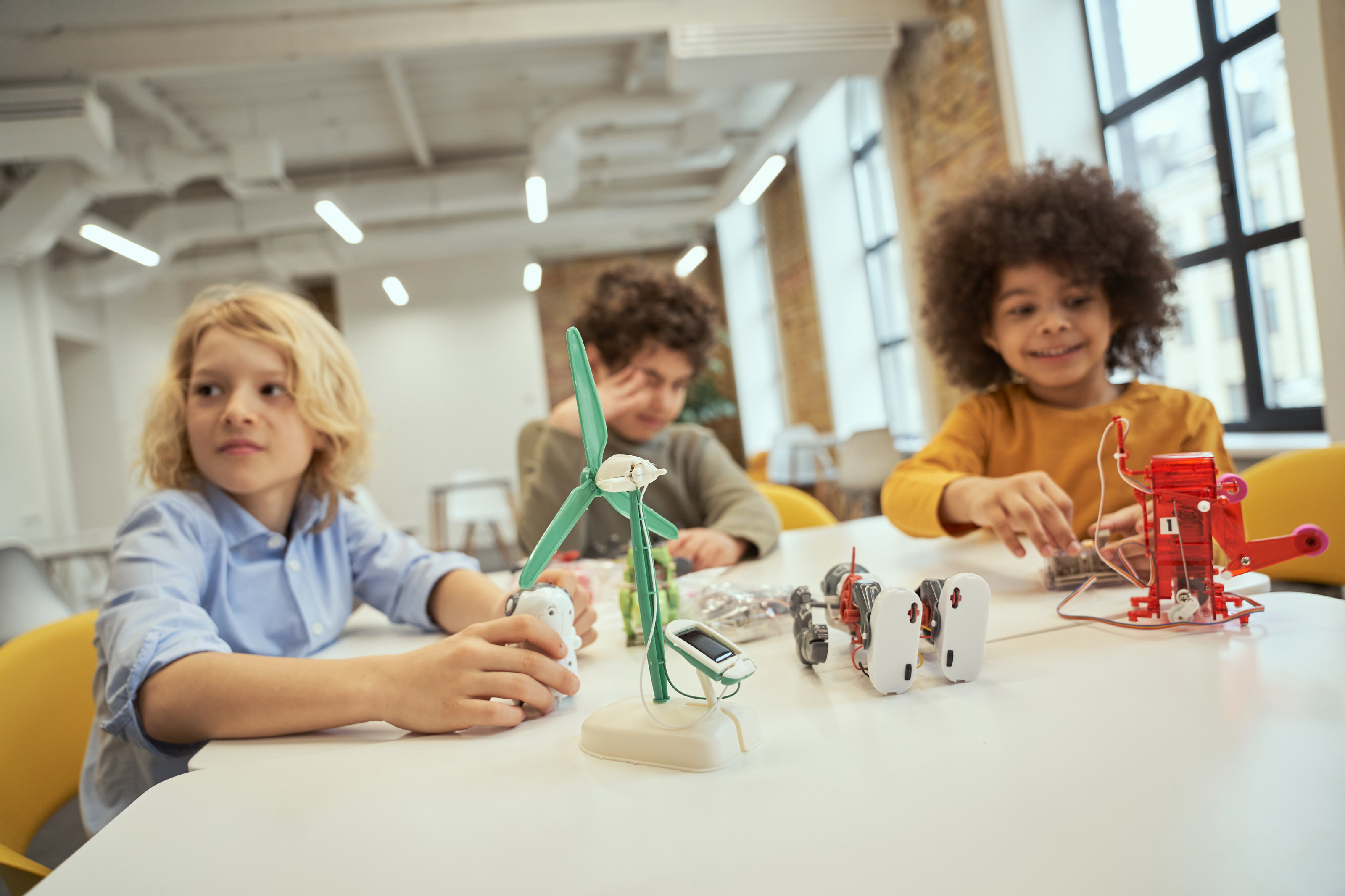 Drei Kinder sitzen an Tisch auf dem Roboter und Modell eines Windrades stehen