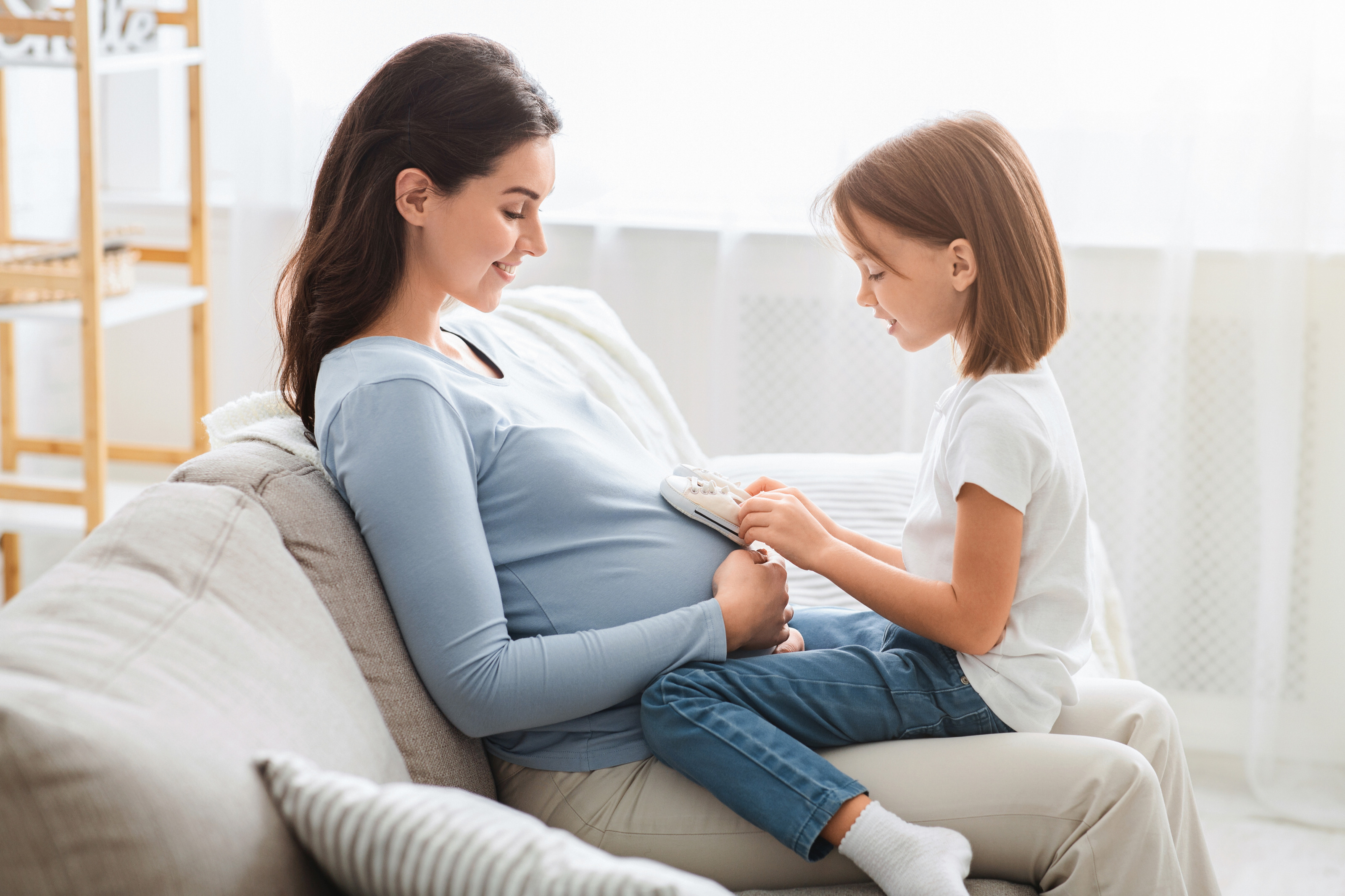 Kind sitzt bei einer schwangeren Person auf der Couch und hält Babyschuhe auf den Bauch