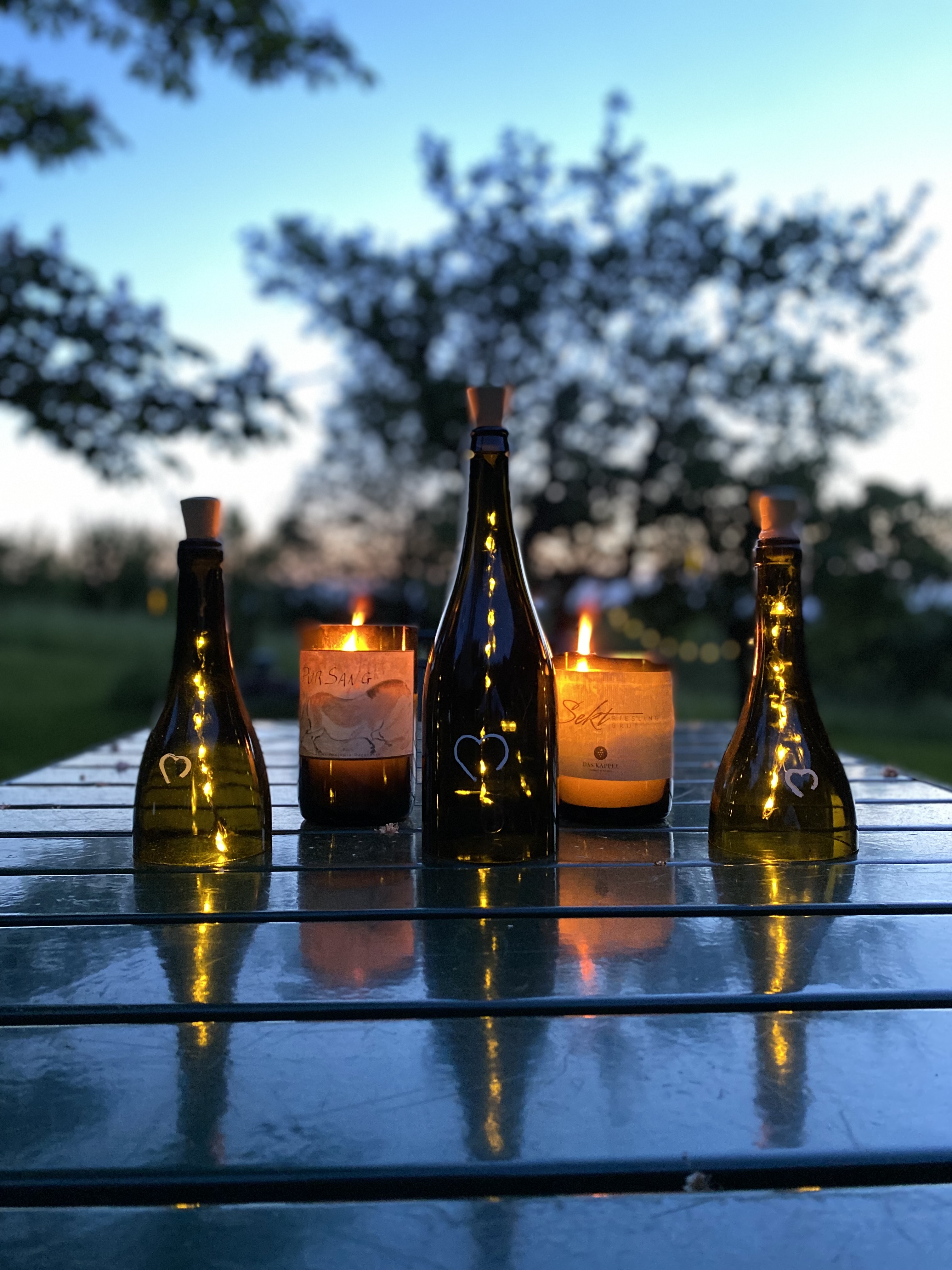 fünf aufgeschnittene Weinflaschen auf einem Tisch, umfunktioniert als zwei Kerzen und drei Solarlampen