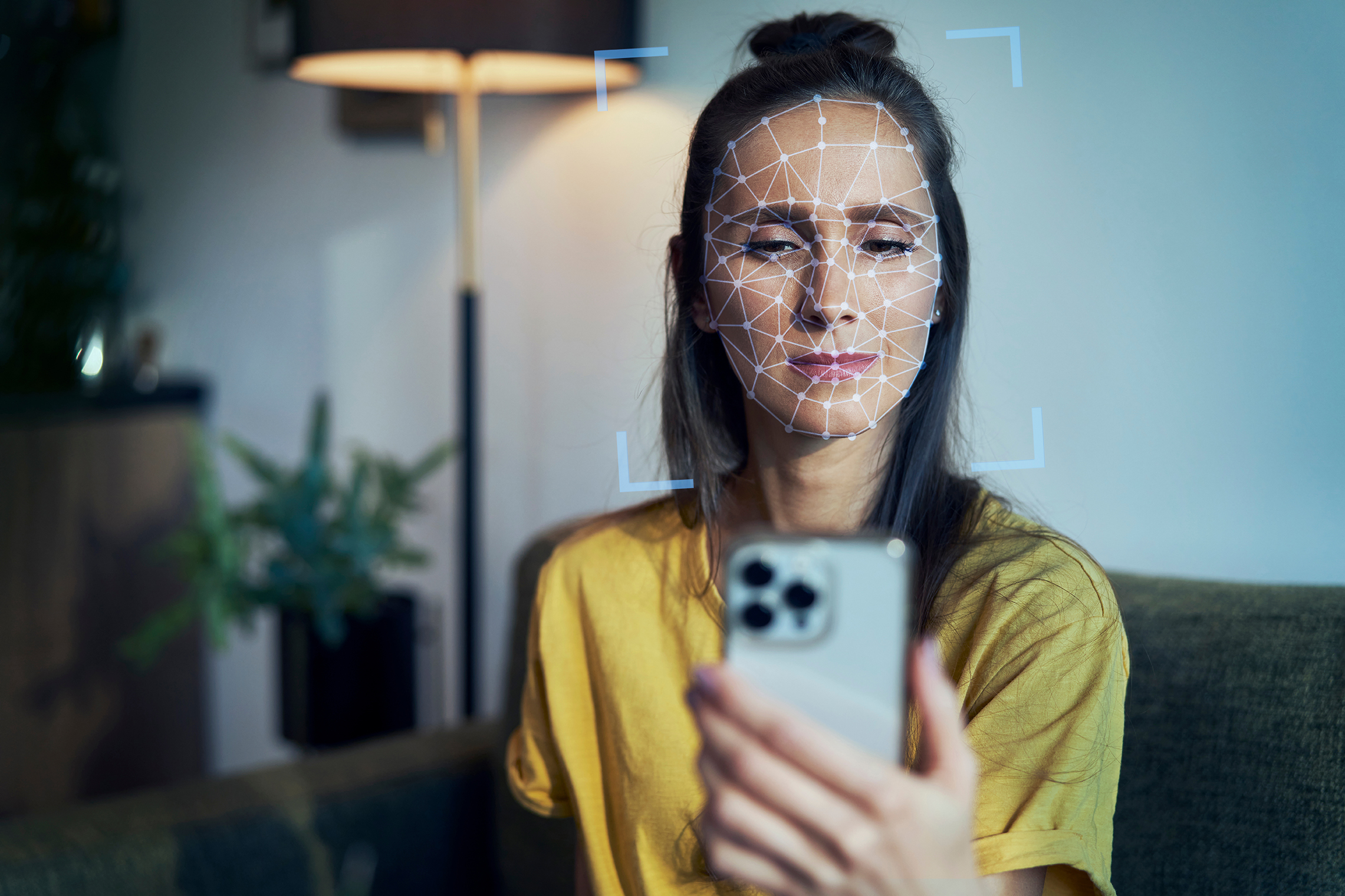 text     Person sitzt auf einer Couch in einem Wohnraum und hält ein Smartphone vor das Gesicht, auf der Gesichtsfläche sowie rund um das Gesicht zeigt sich eine Vermessung als Visualisierung der Gesichtserkennung