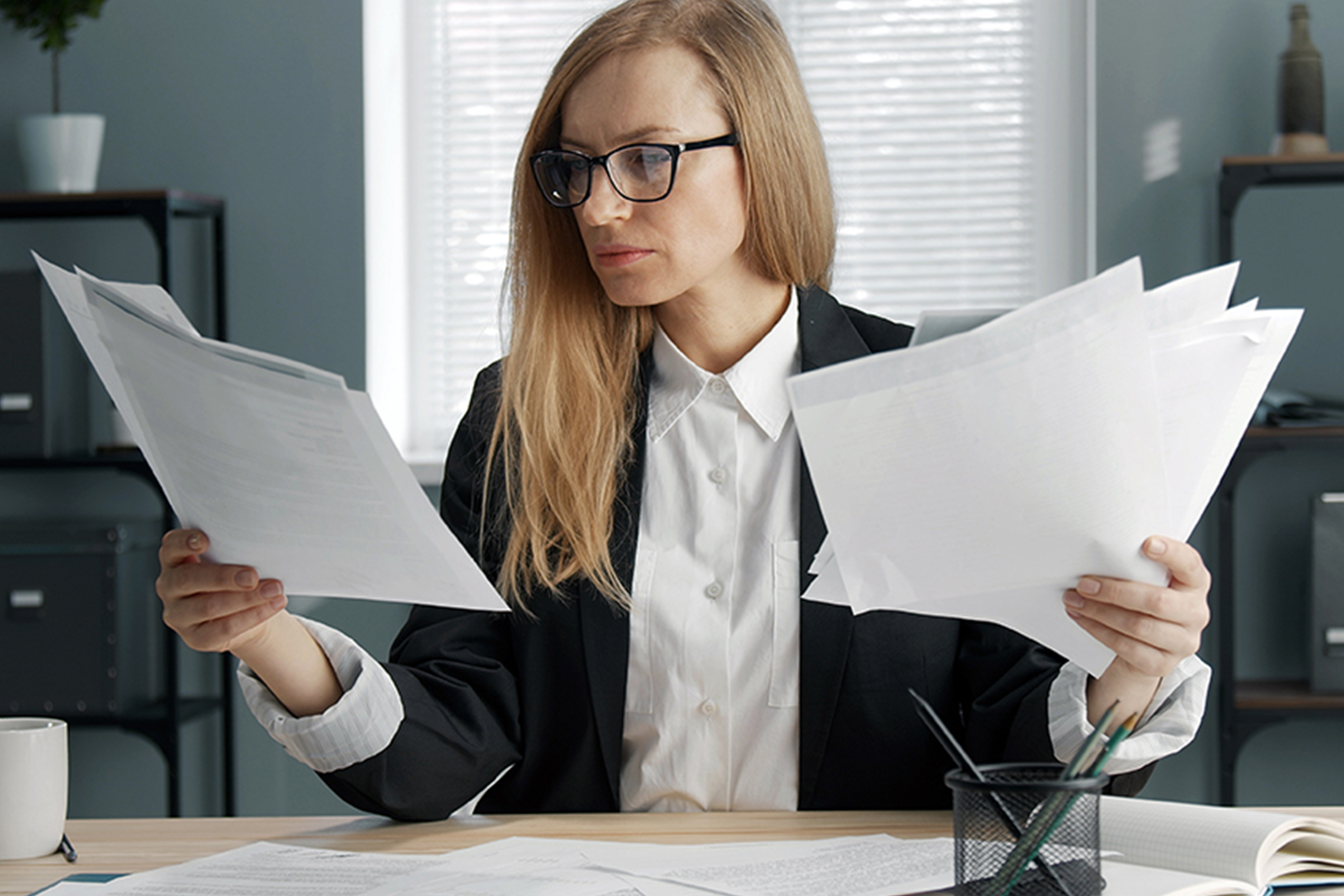 Concentrated blond business woman sitting at office desk holding heaps of documents, paper reports