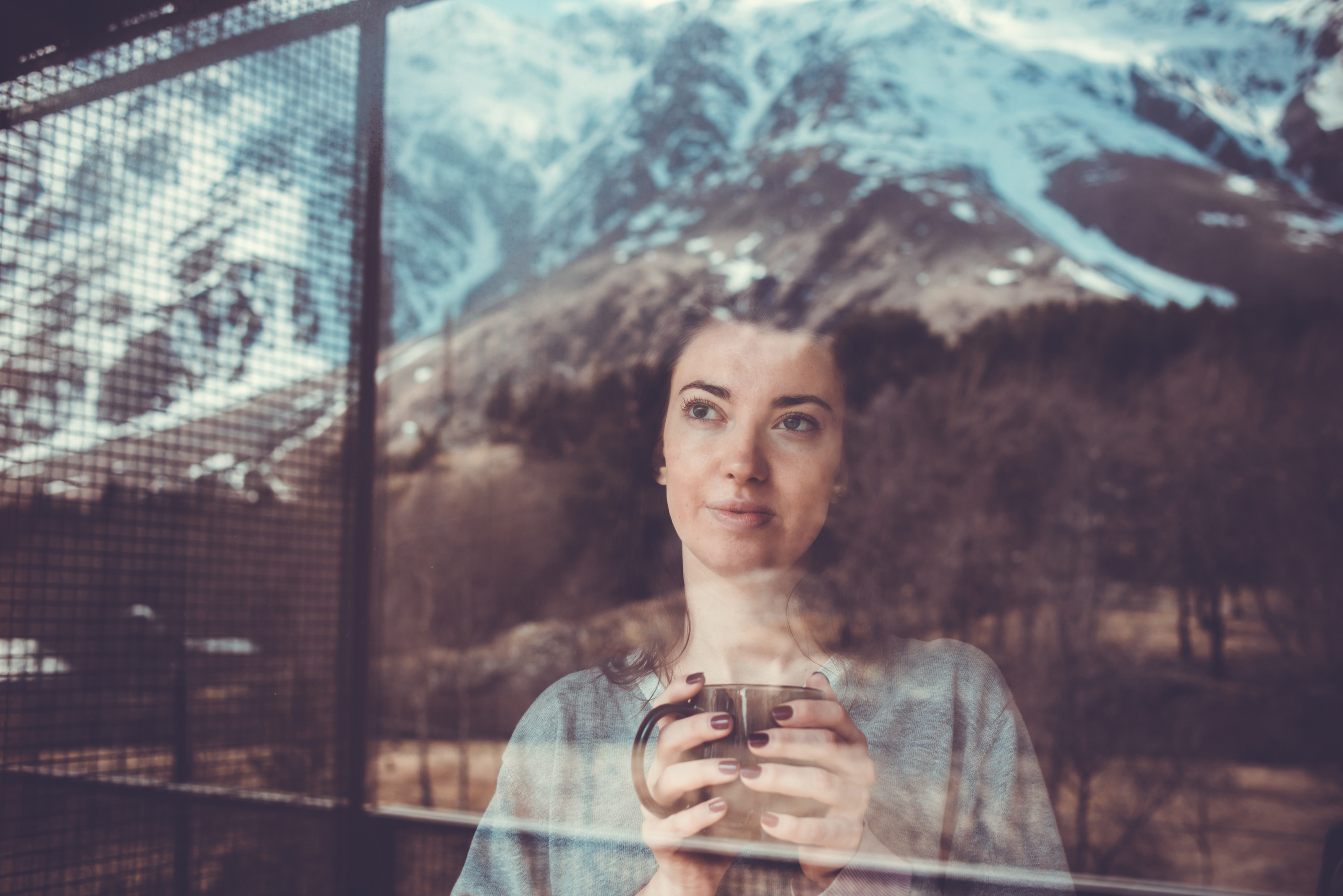 Person hält Tasse in beiden Händen und blickt durch Fenster auf Bergpanorama, das sich in diesem spiegelt