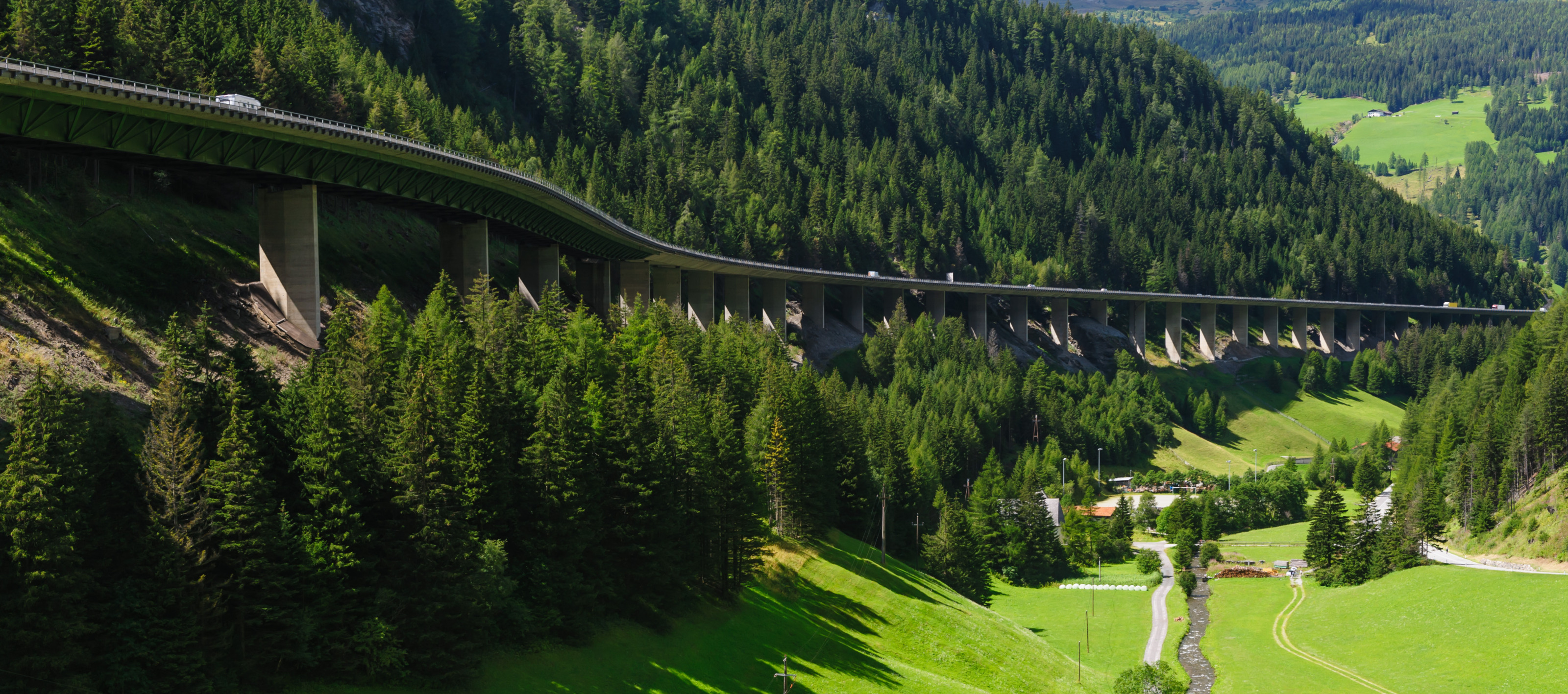 In der rechten Bildhälfte ist eine grüne Wiese mit grünen Bäumen. In der linken Bildhälfte führt eine Straße durch die Landschaft. Die Straße steht auf Betonpflöcken. Links neben der Straße ist ein ansteigender Hügel mit grünen Bäumen